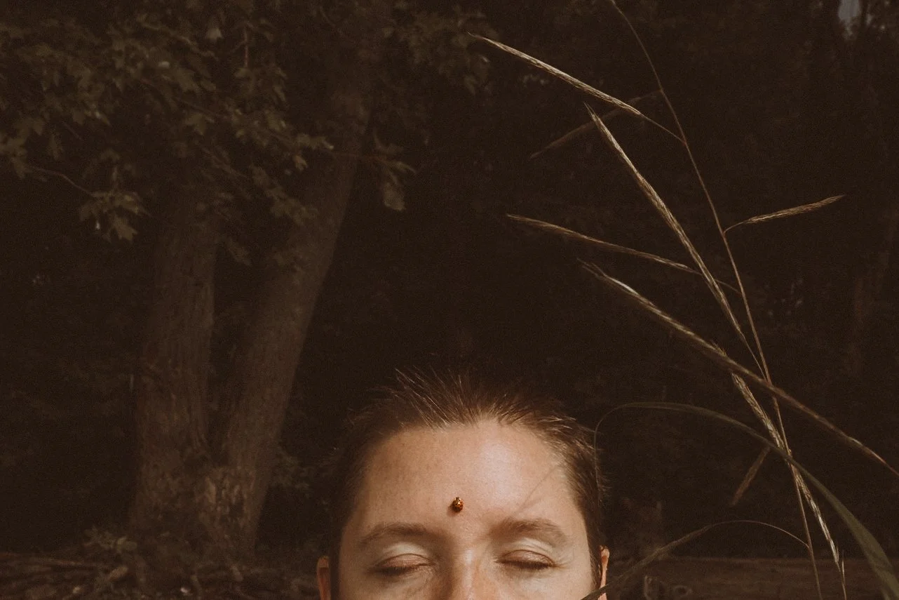 A person with closed eyes and a bindi on the forehead outdoors at night, with trees and tall grass visible in the dark background.