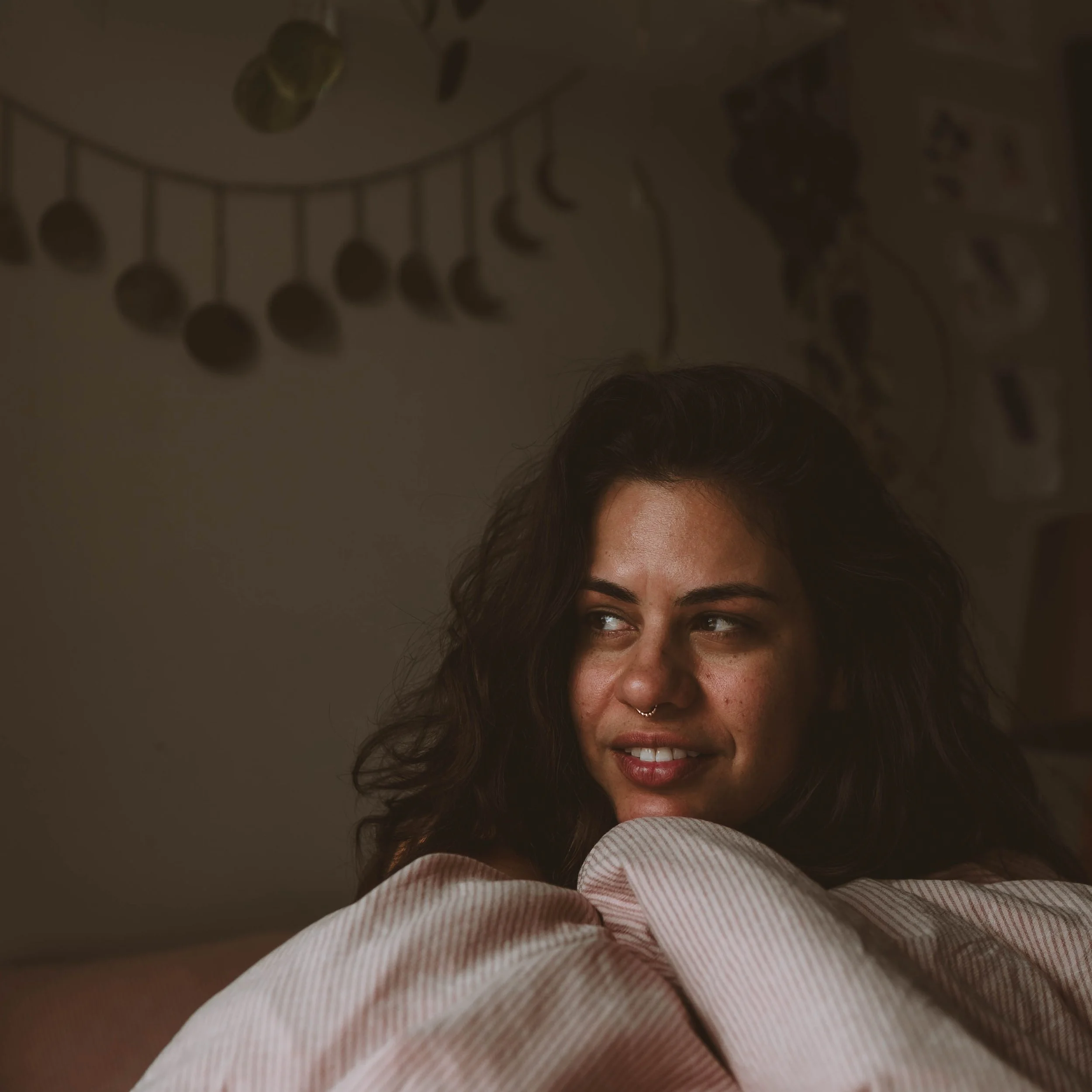 A woman with dark, curly hair smiling and looking to the side, with a septum piercing, sitting in a dimly lit room with decorative items on the wall behind her.