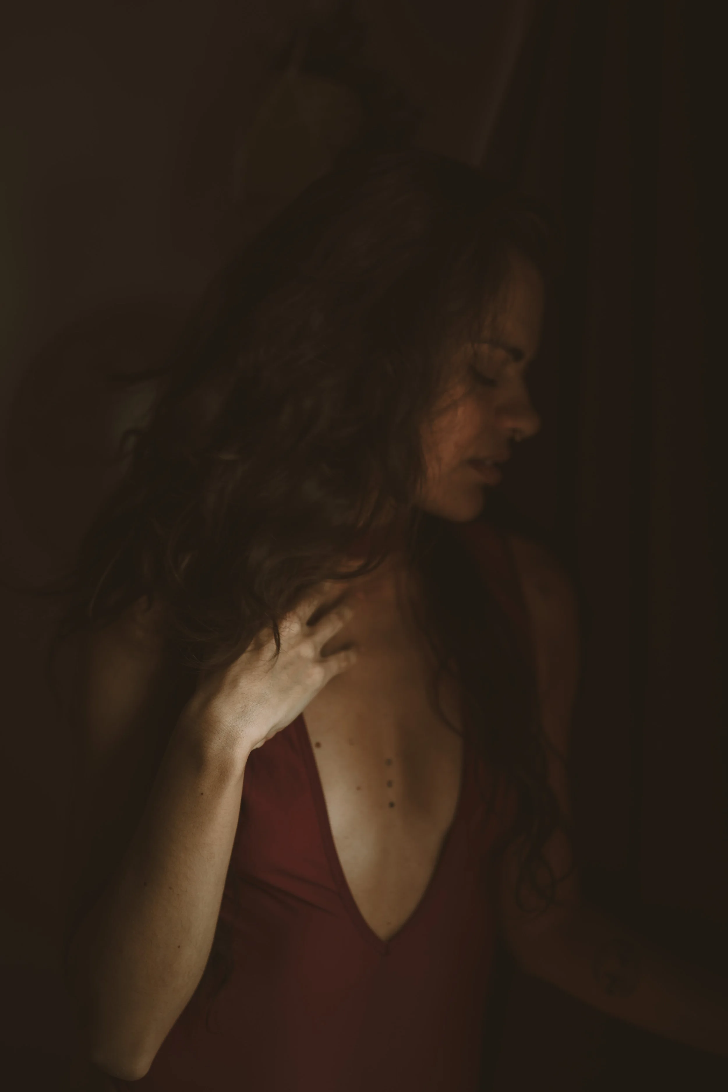 A woman with long brown hair wearing a red dress, with her hand on her chest and their head tilted down, in a dimly lit setting.