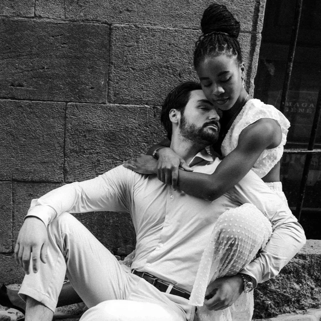 A black and white photo of a man sitting on a bench, with a woman embracing him from behind. The man has dark hair and a beard, and is wearing a light-colored shirt and pants. The woman has her hair styled in a high bun and is wearing a sleeveless, textured dress. They are sitting close against a stone wall.