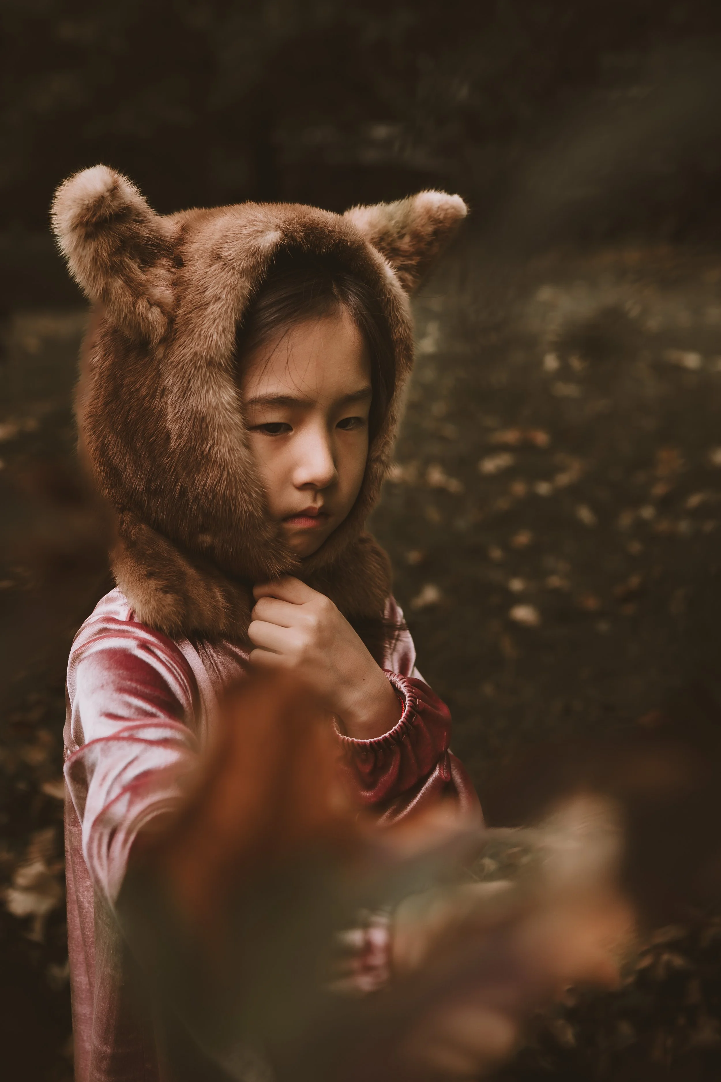 A young girl with dark hair appears contemplative as she wears a fox costume hood with realistic fur and ears. She is outdoors with a blurred background of trees and fallen leaves.