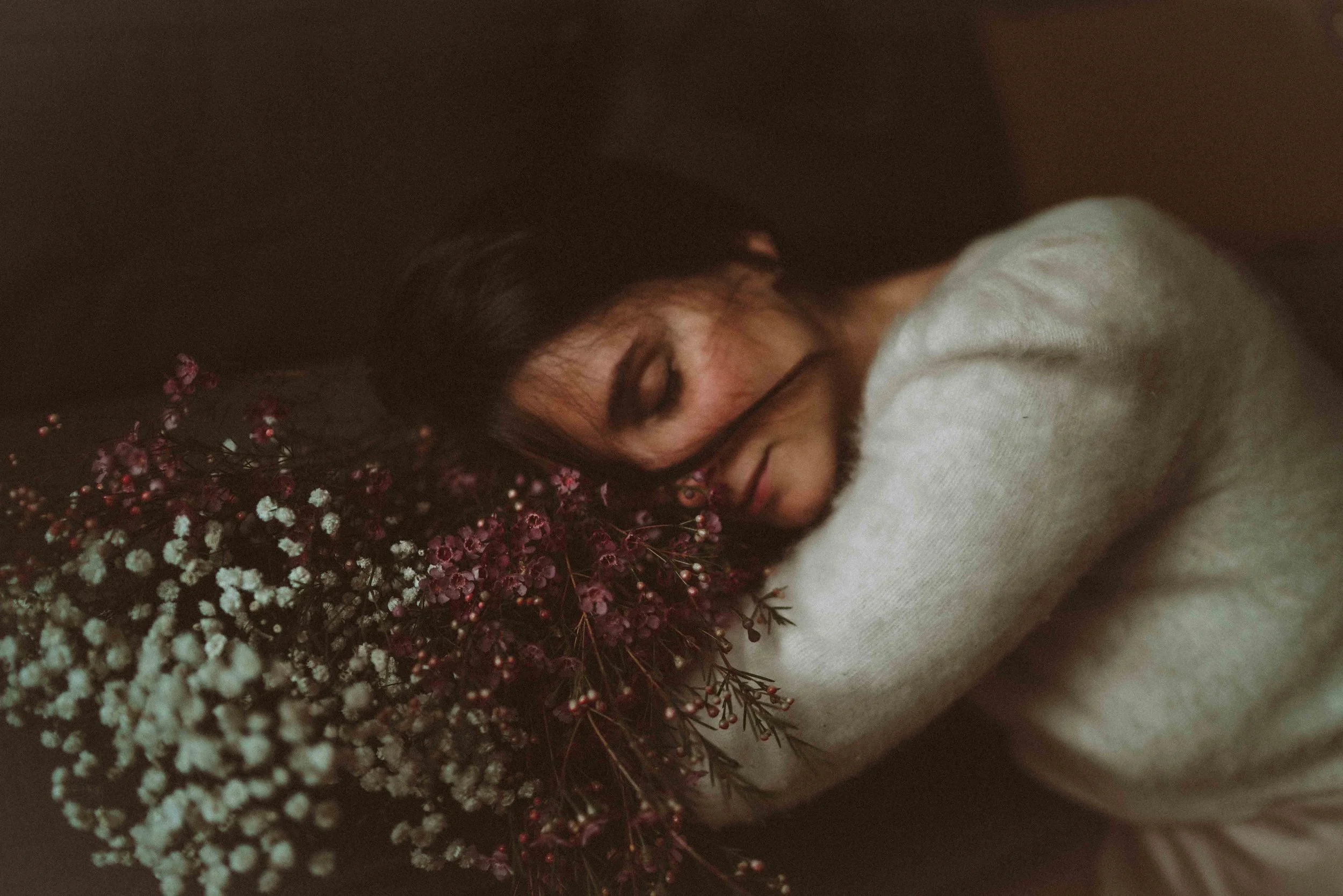 A woman with dark hair and glasses, wearing a light-colored sweater, rests her head on a dark background, surrounded by a bouquet of small purple, white, and pink flowers.
