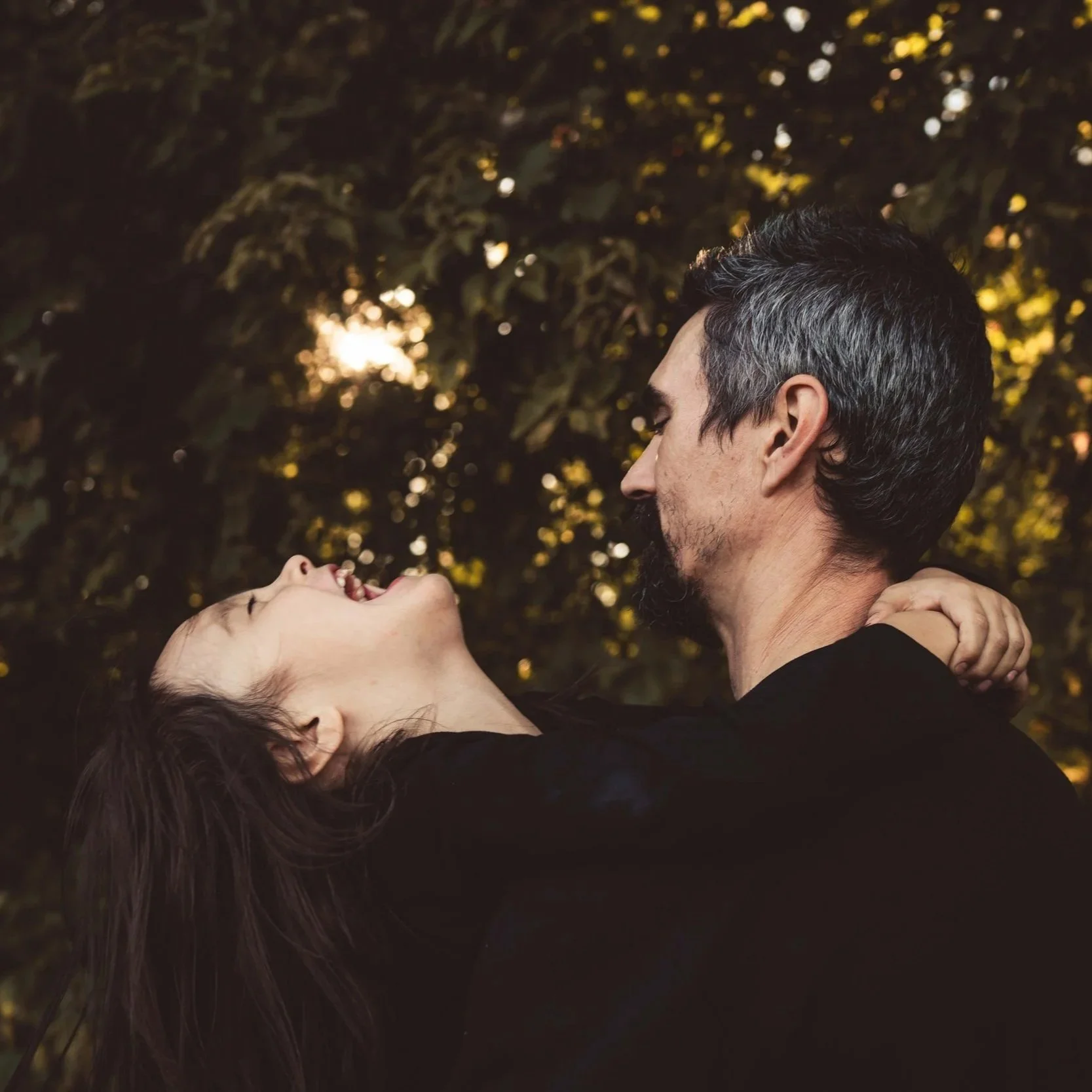 A man with dark hair and a beard holding a woman with long dark hair, both with their eyes closed, outdoors during sunset, with trees and golden light in the background.