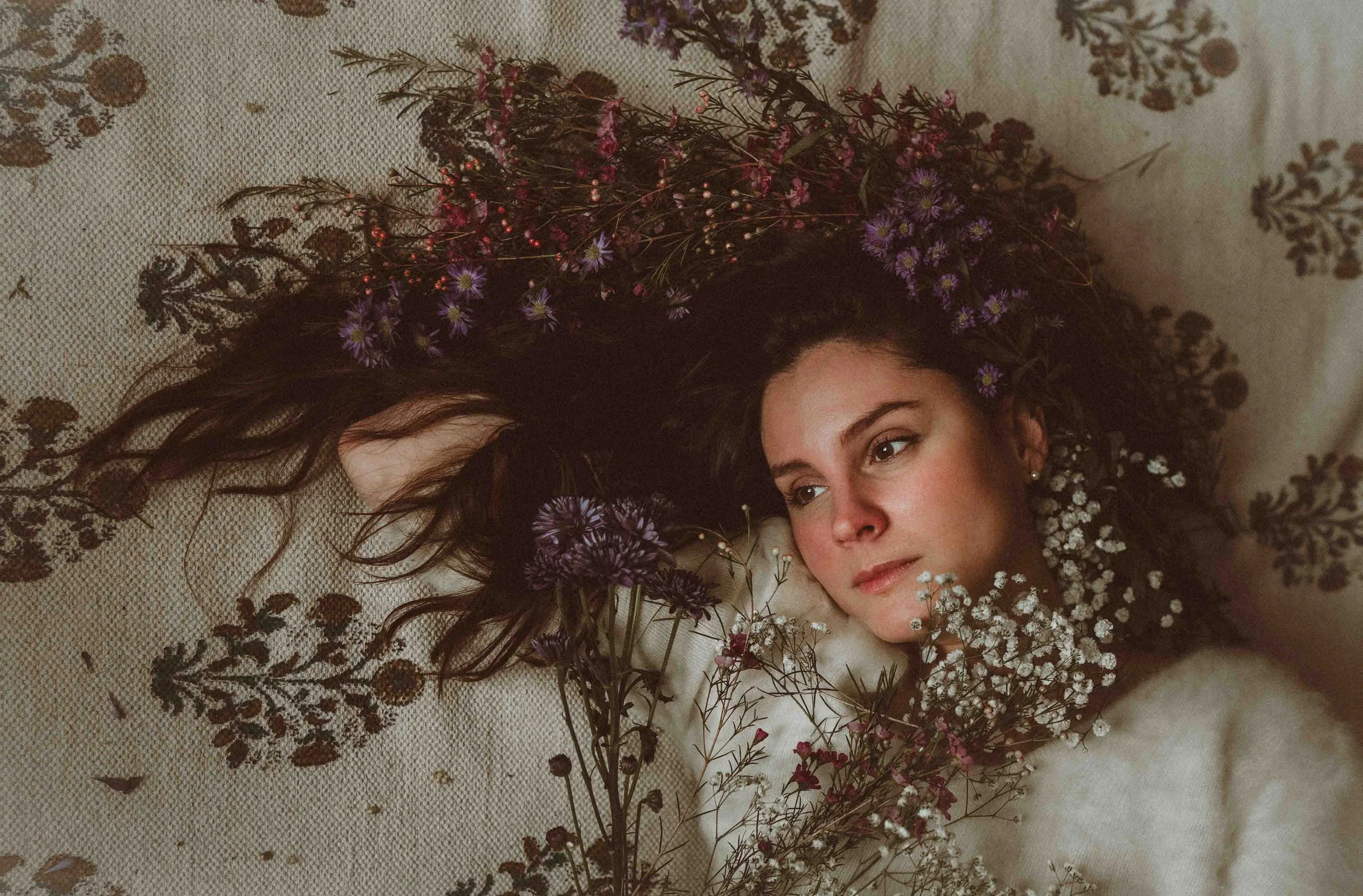 A woman with long dark hair lying on a patterned surface surrounded by purple and white flowers.