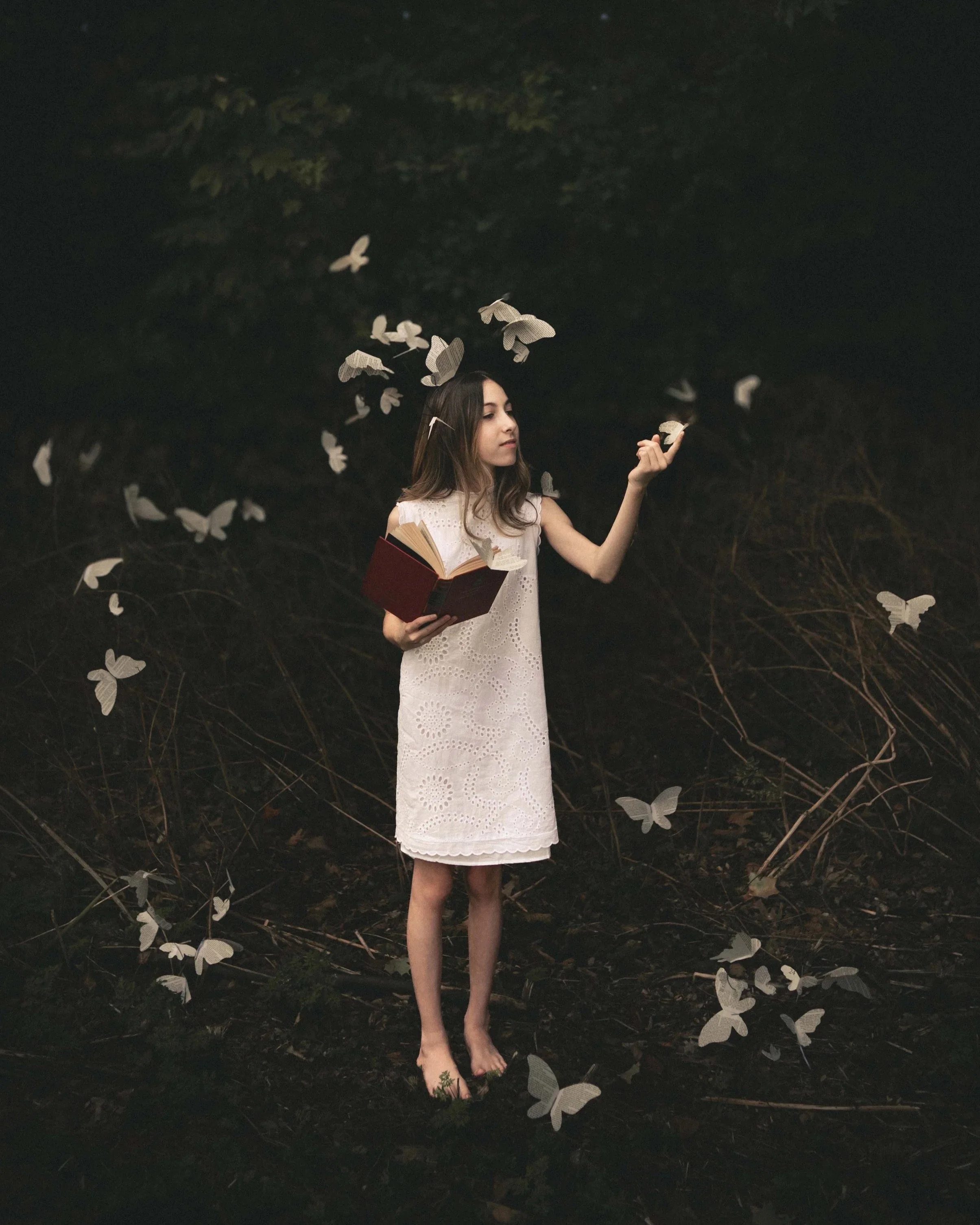 A young woman with long brown hair stands outdoors in the dark, holding an open book. She is wearing a white, eyelet lace dress and appears to be gently touching a paper butterfly with her right hand. Several paper butterflies are flying around her, 