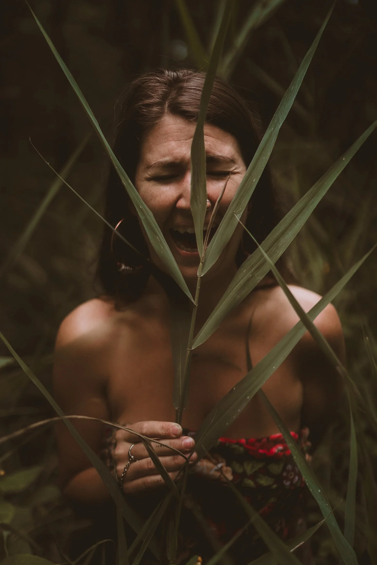 A woman with a big smile, squinting her eyes, is sitting amongst tall grass, partly obscured by the greenery, with her hand visible in front of her chest.