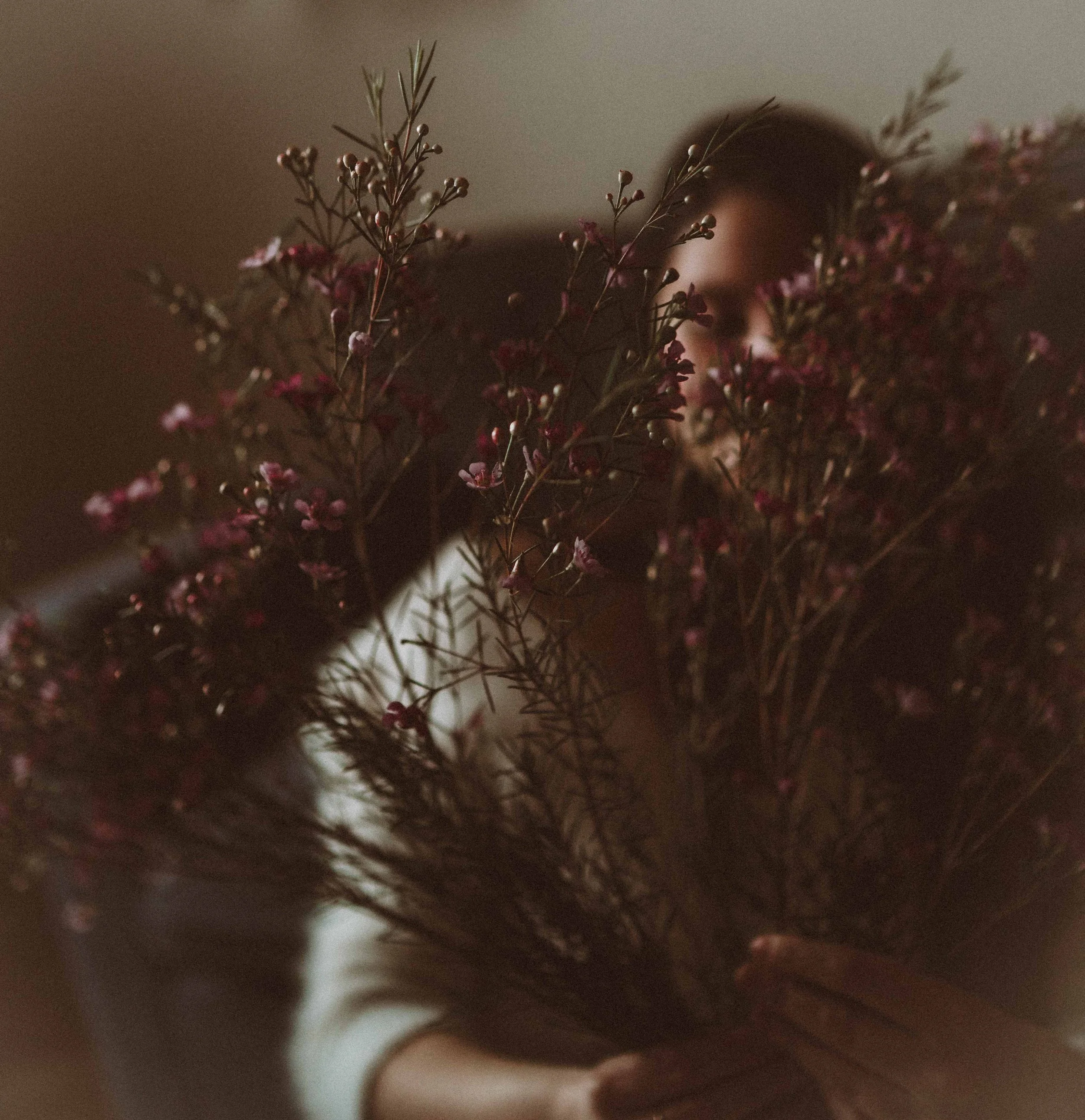 Person holding a bouquet of pink and purple flowers with dark leaves, partially obscuring their face in a dimly lit setting.