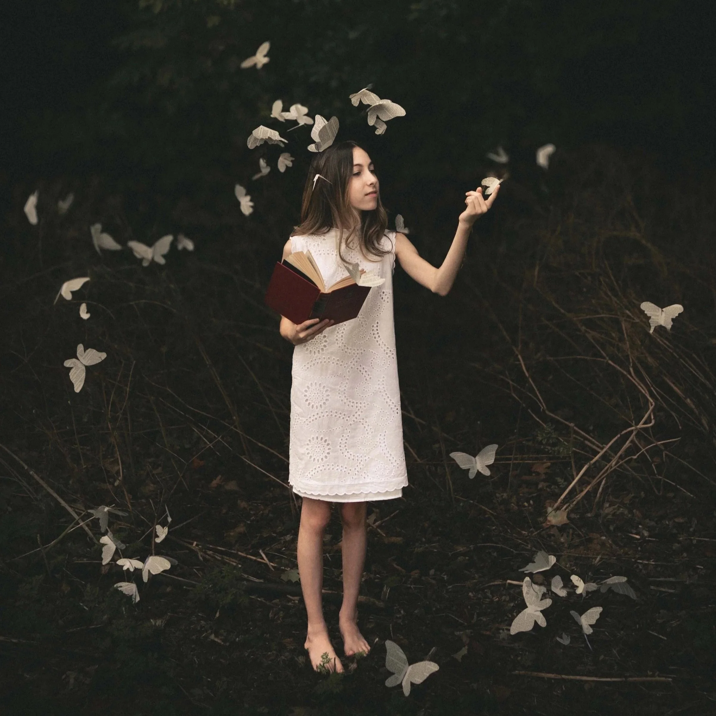 A woman in a white dress is standing outdoors at night, holding an open book and surrounded by white butterflies fluttering around her.