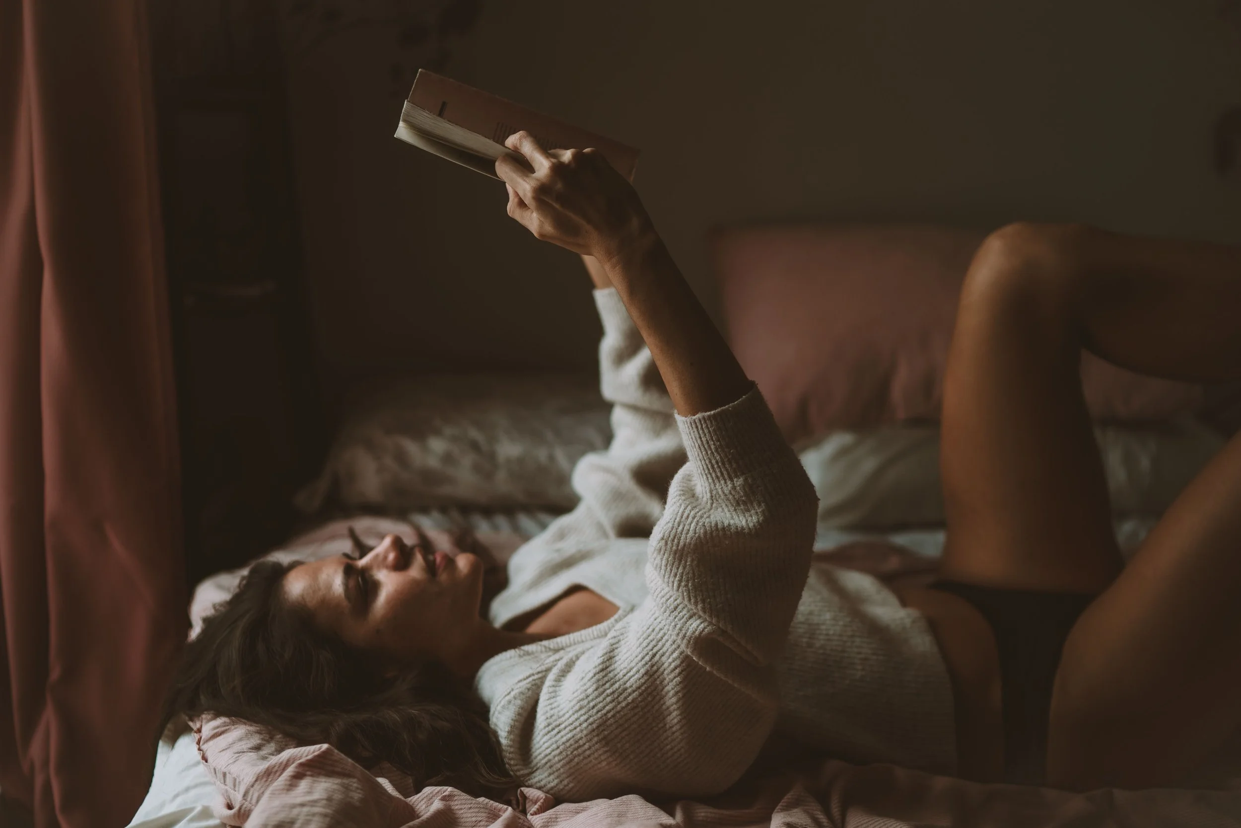 A woman lying in bed reading a book, wearing a sweater, in a dimly lit room.