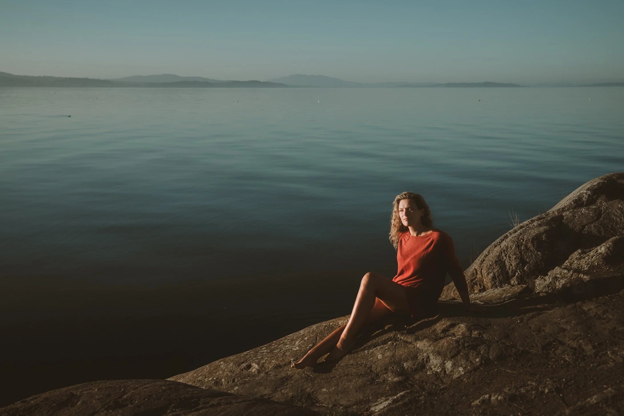 A woman with curly hair wearing a red dress sitting on rocks by a calm body of water, with mountains in the distance on a clear day.