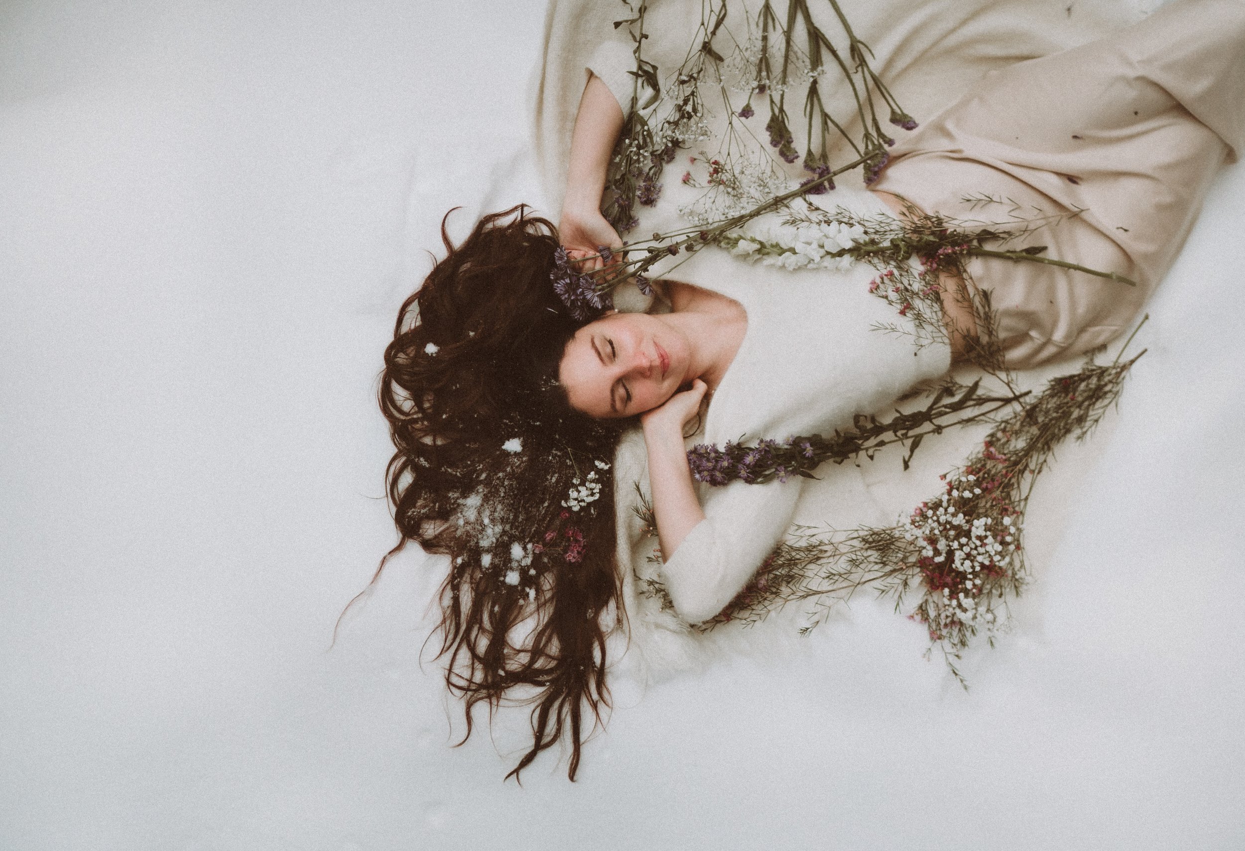 A woman lying on a white surface surrounded by dried flowers, with her eyes closed and holding a flower.