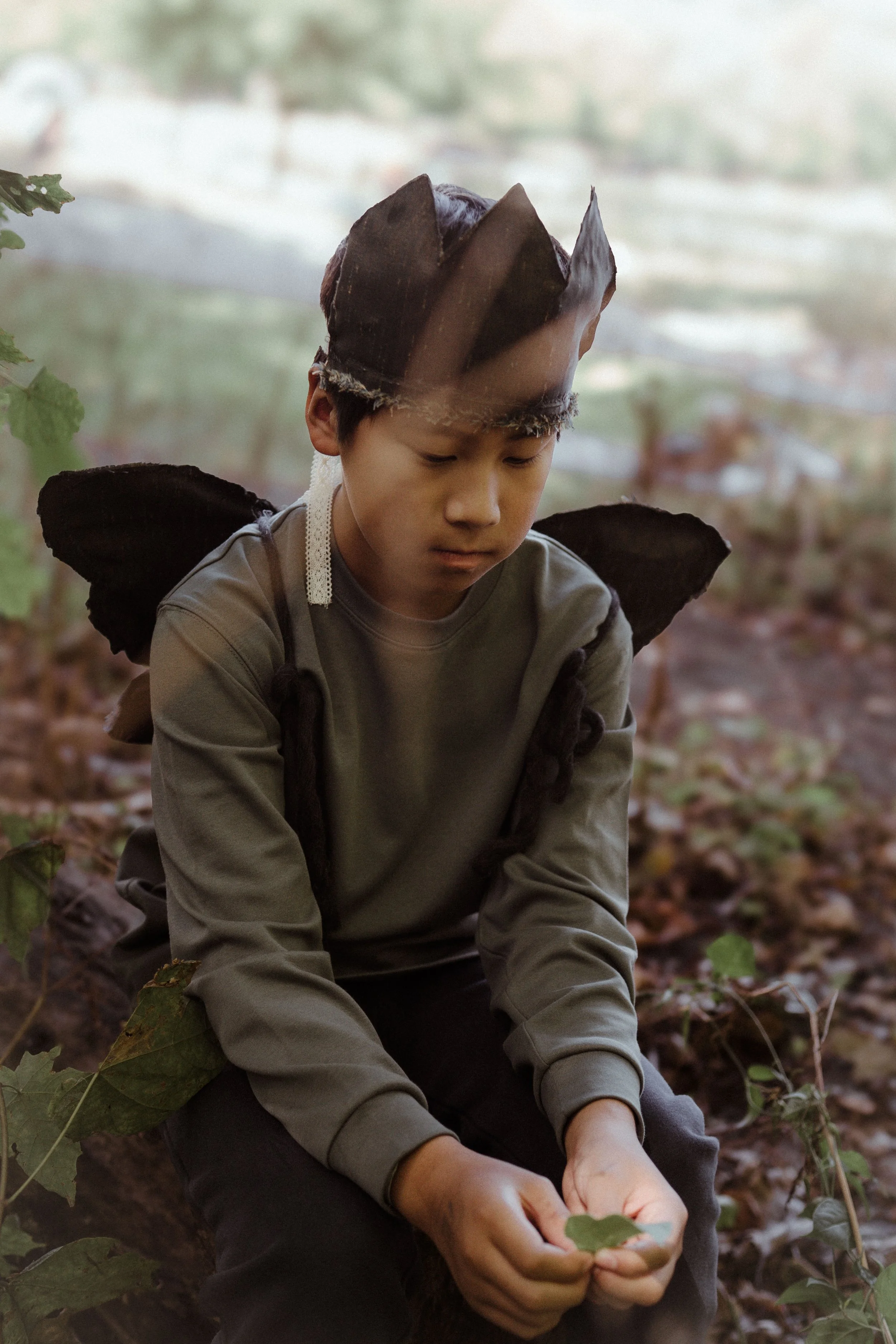 A young boy dressed as a fairy, wearing a brown paper crown and black wings, is sitting outdoors among fallen leaves, holding a green leaf in his hands and looking down thoughtfully.