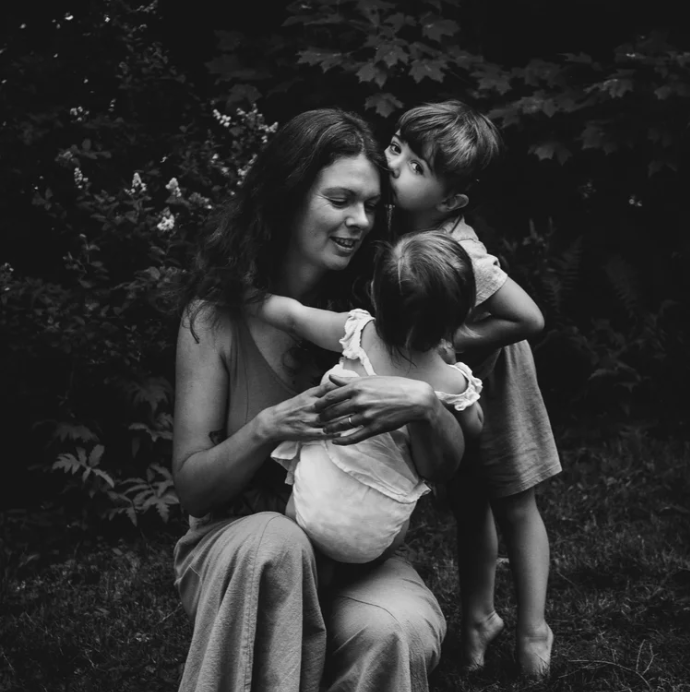 A woman lying on a bed of ferns with her legs extended and one arm behind her head, wearing a beige dress with embroidery.