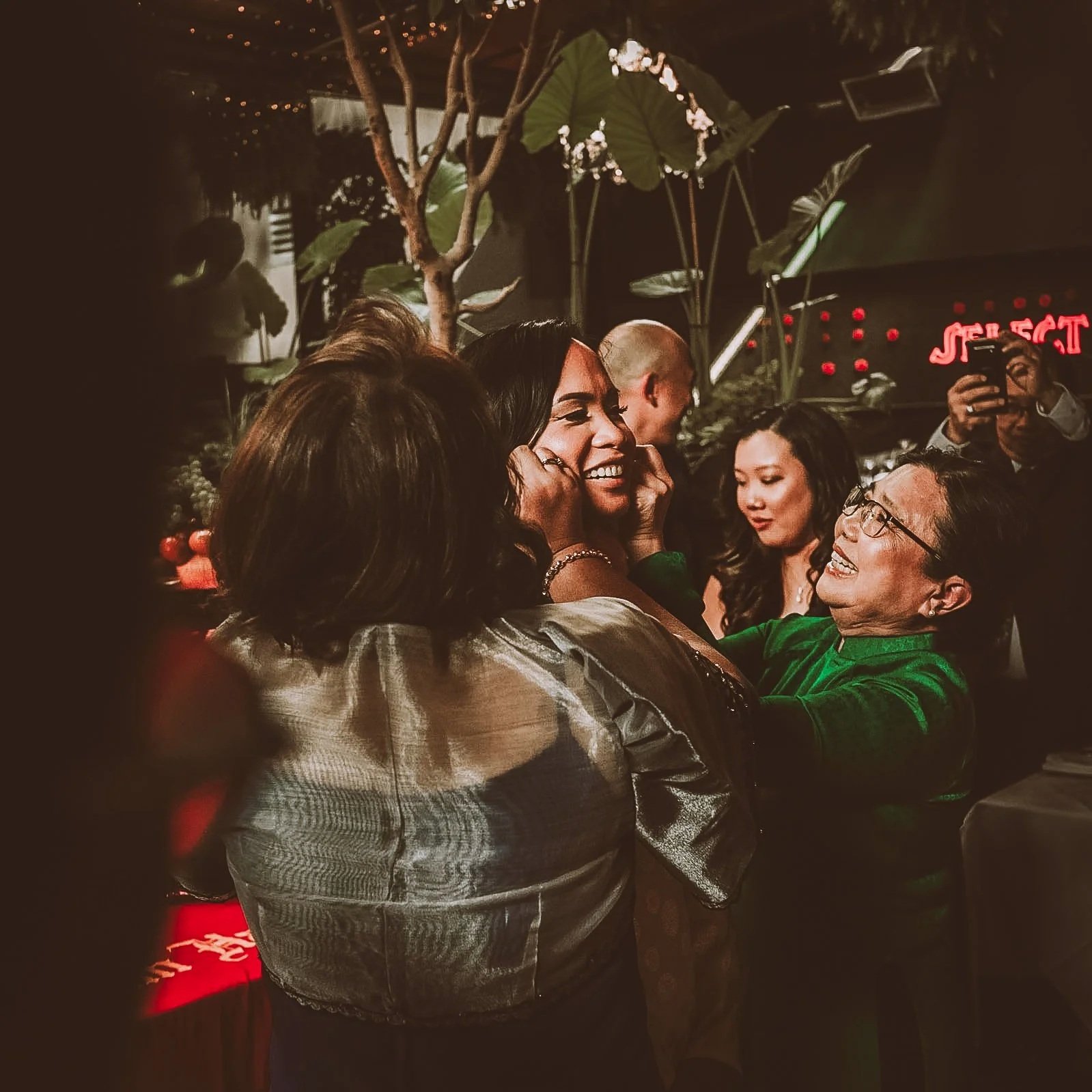 People at a social gathering, smiling and engaging in conversation in a warmly lit environment with plants and decorative lights in the background.