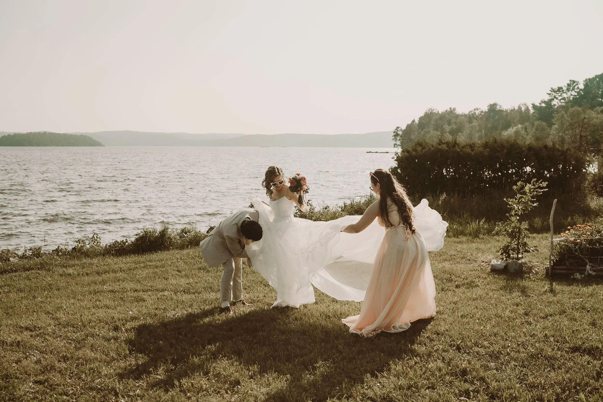 A bride in a white wedding dress holding a bouquet stands on a grassy lakeside, assisted by a woman in a light pink dress, with a man in a light suit bending over beside her. The scene is bright and sunny, with a large body of water and trees in the 