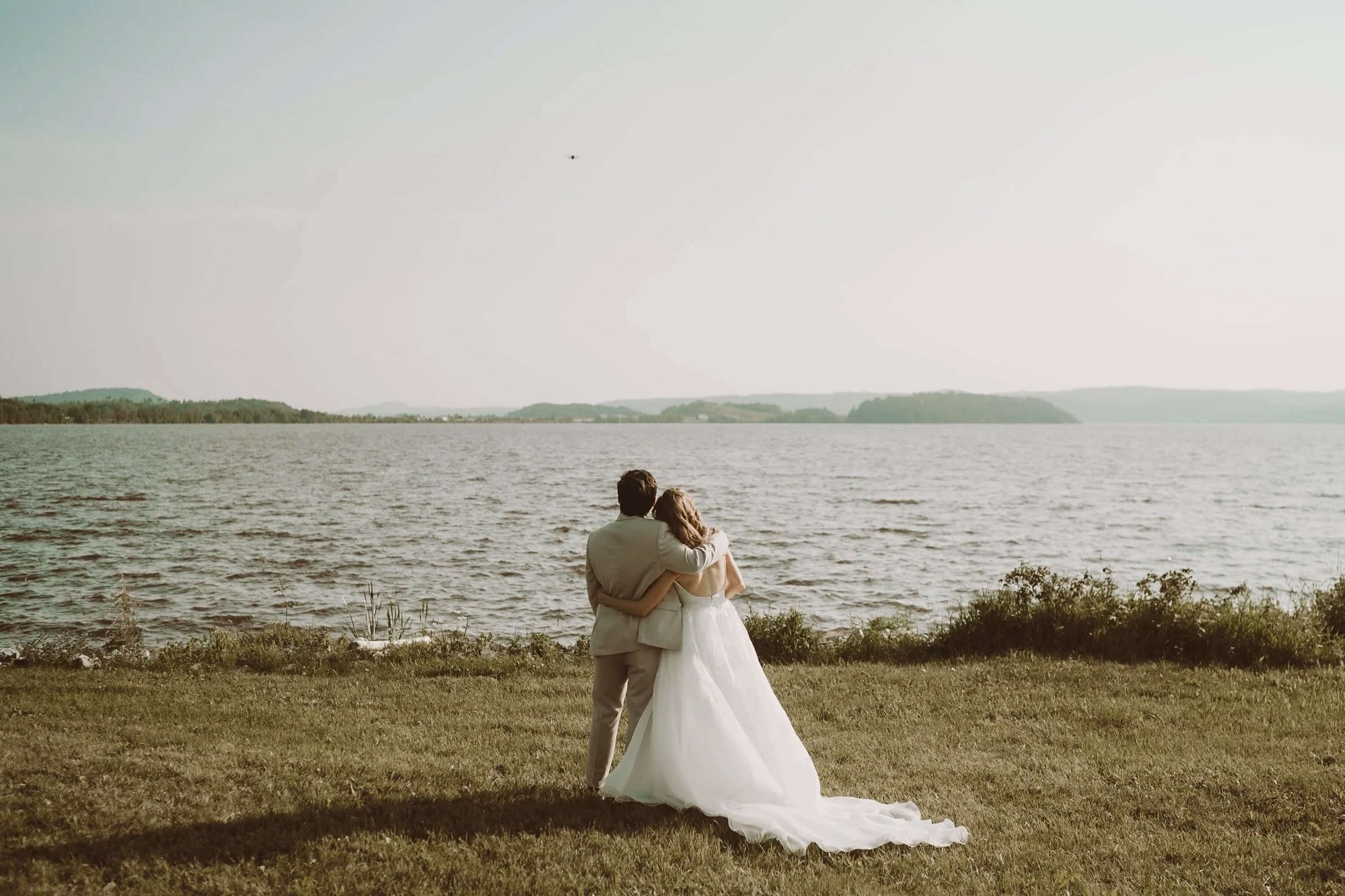 A bride and groom standing close together by a lake, with the bride in a white wedding dress and the groom in a light-colored suit, embracing each other and looking at the water.