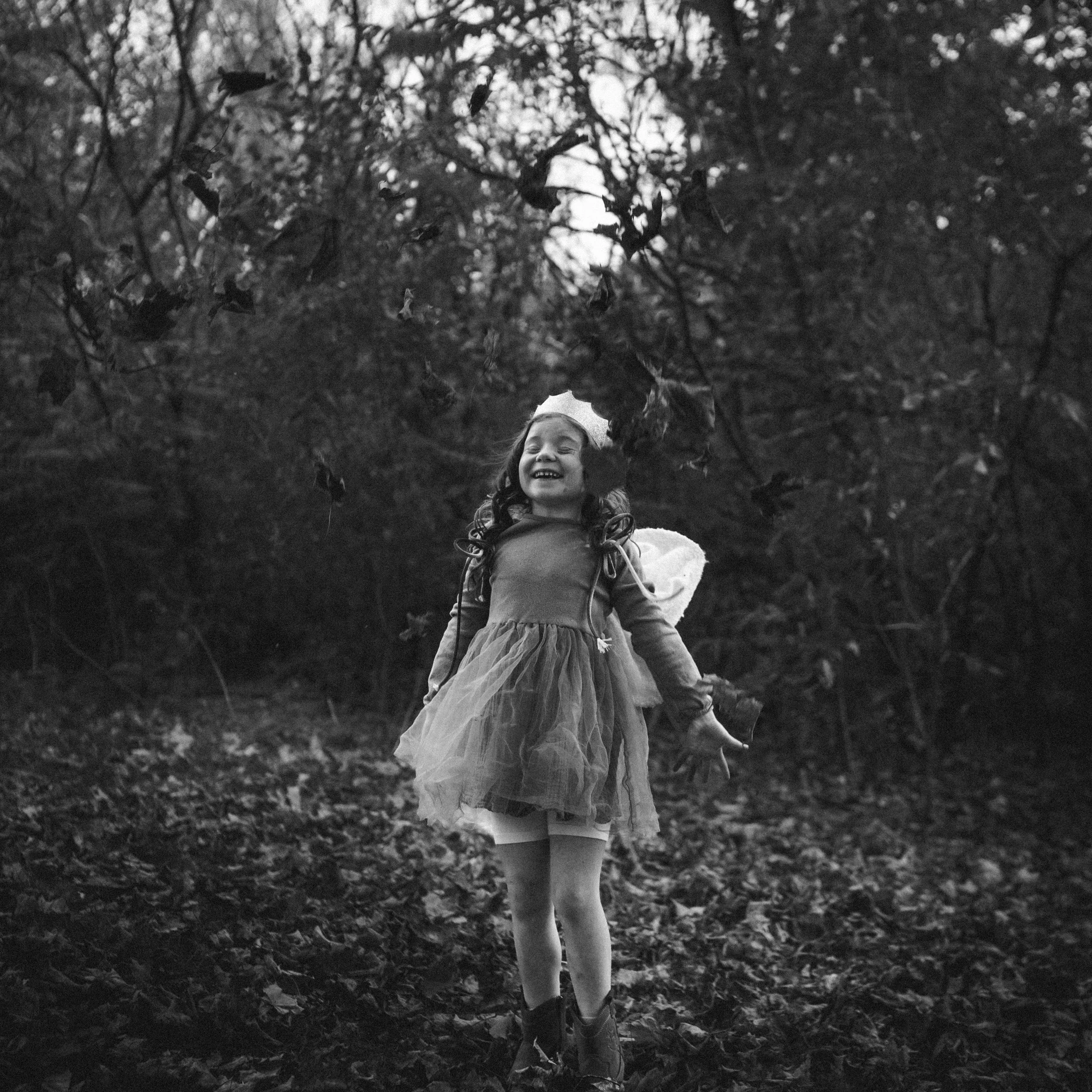 A black and white photo of a girl outdoors, smiling, standing among trees and fallen leaves, with a large pile of leaves above her.