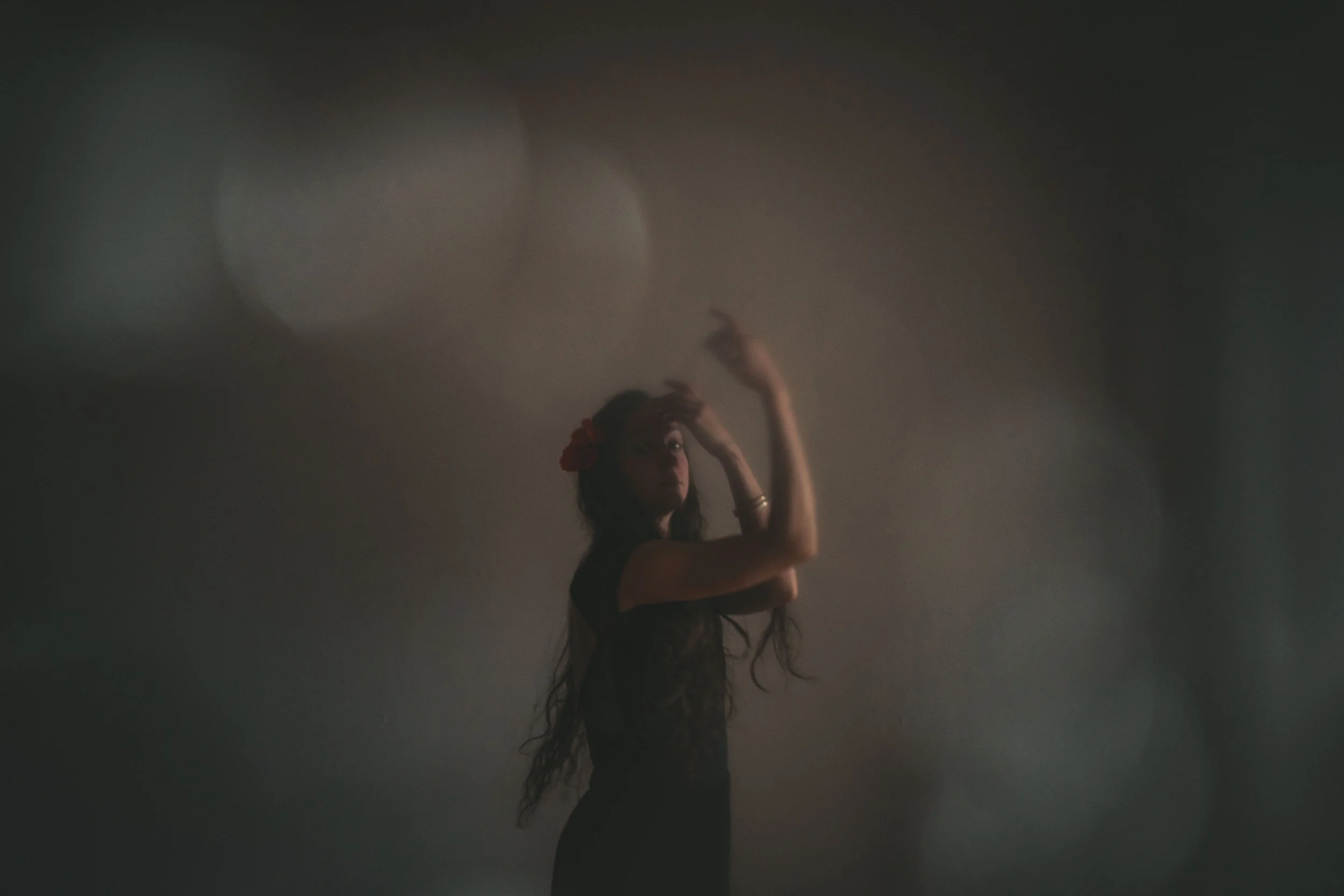 A woman with long curly hair, wearing a black dress and a red flower in her hair, standing behind a foggy or smoky window and gazing outside, with her hands raised near her head.