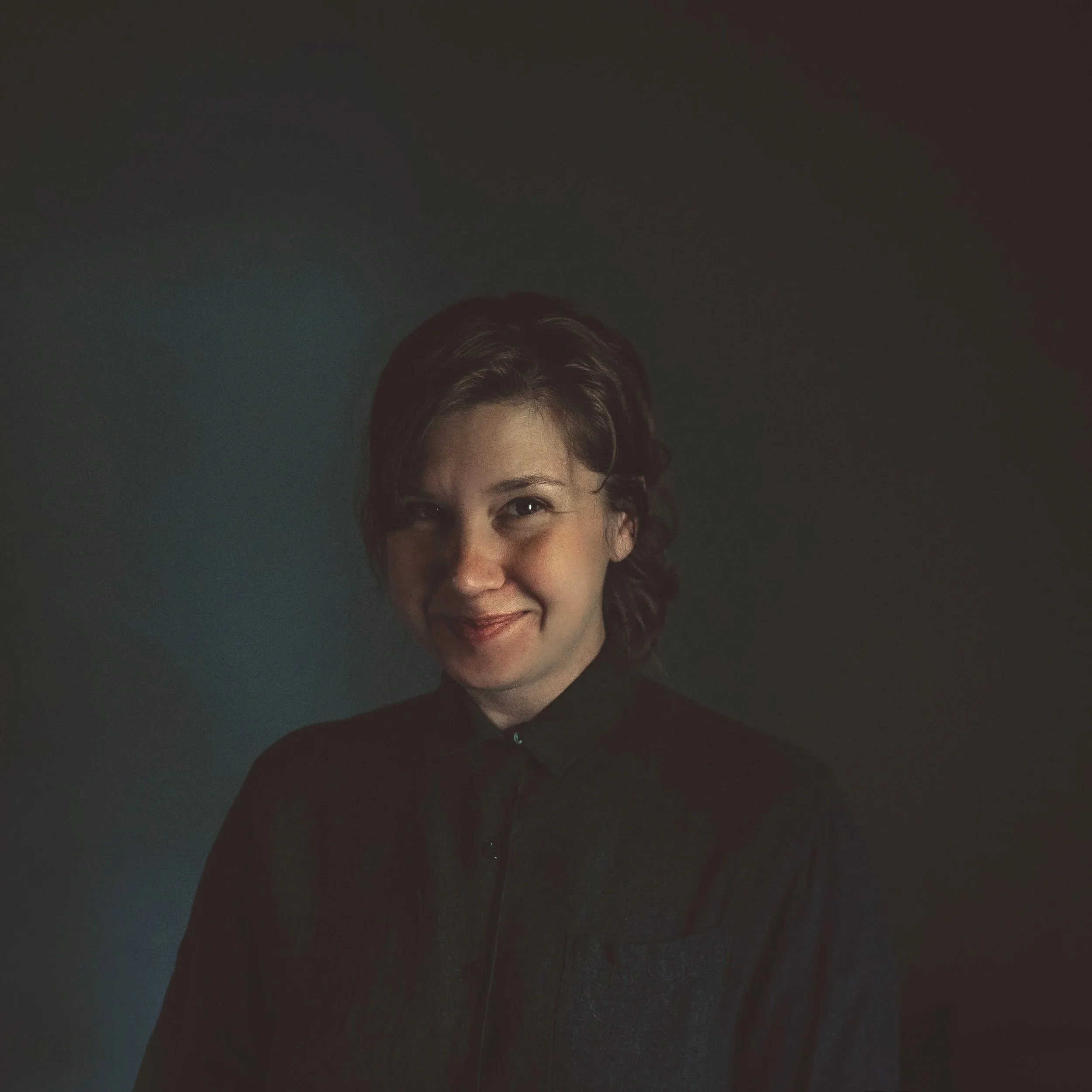 A woman with short wavy brown hair wearing a dark shirt, smiling softly, standing against a dark background with subtle lighting highlighting her face.