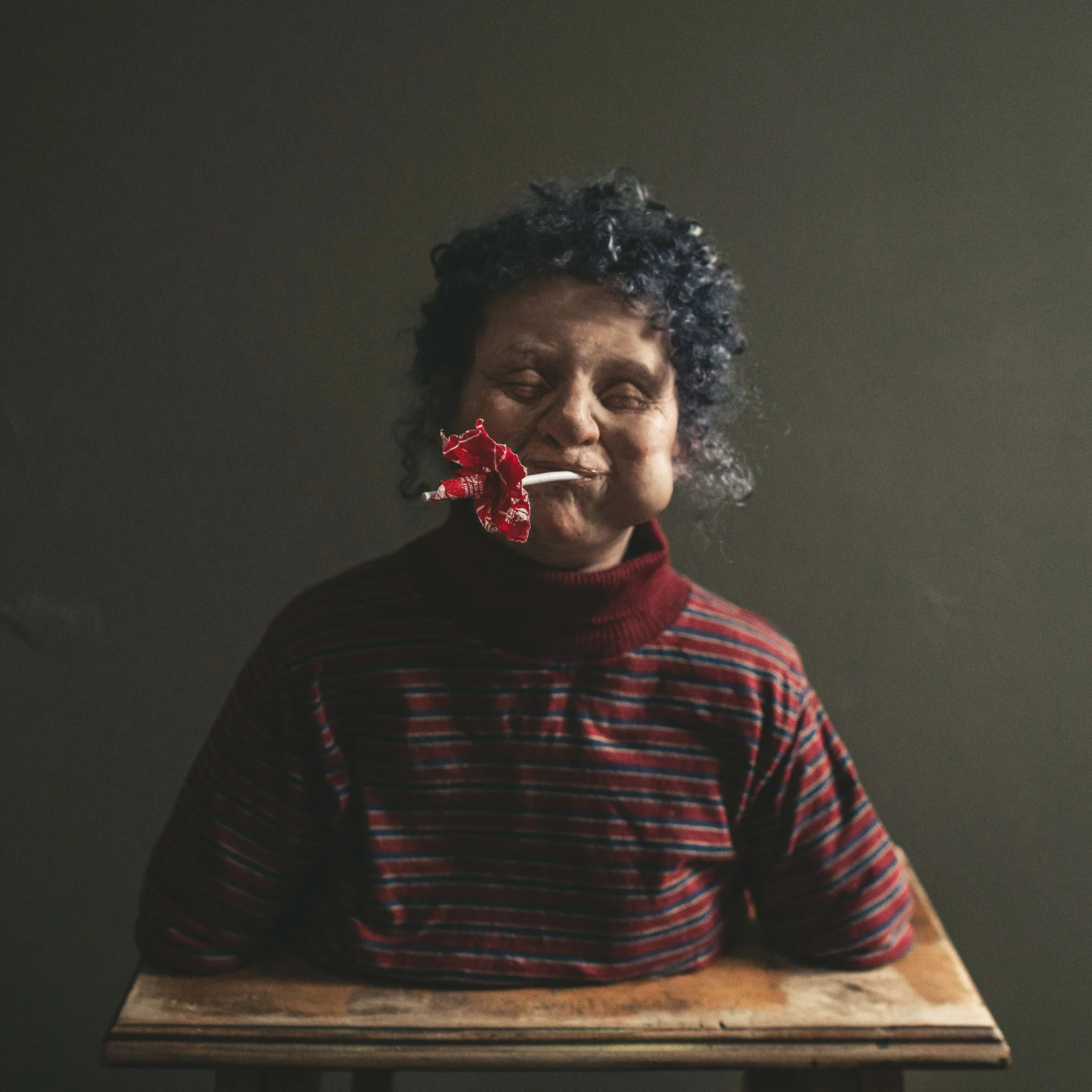 A sculpture of a woman with curly dark hair, wearing a striped red and black sweater, sitting behind a wooden table. She has a playful expression with a chew item in her mouth with a red cloth attached.