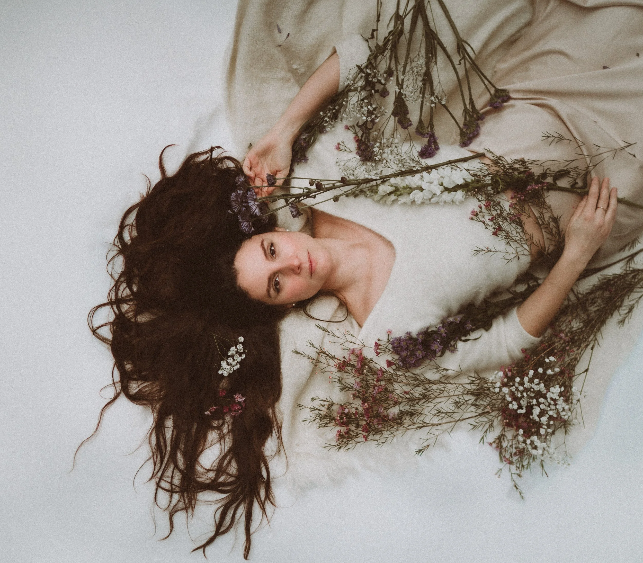A woman with long, dark hair lies on a white surface surrounded by various dried flowers. She is wearing a cream-colored sweater, and her gaze is directed at the camera.