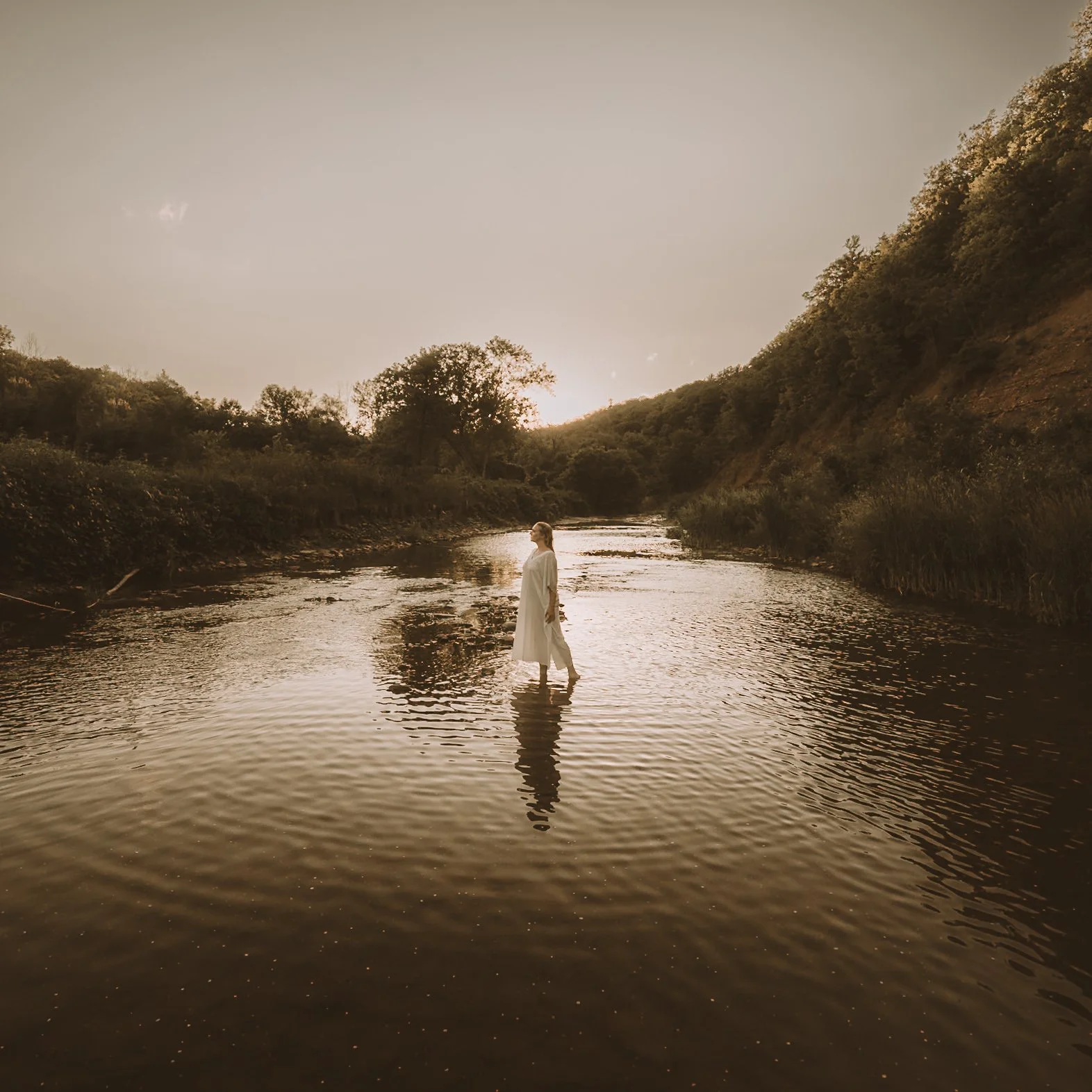 A person wearing a white dress standing in a calm river with trees and hills in the background at sunset or sunrise.