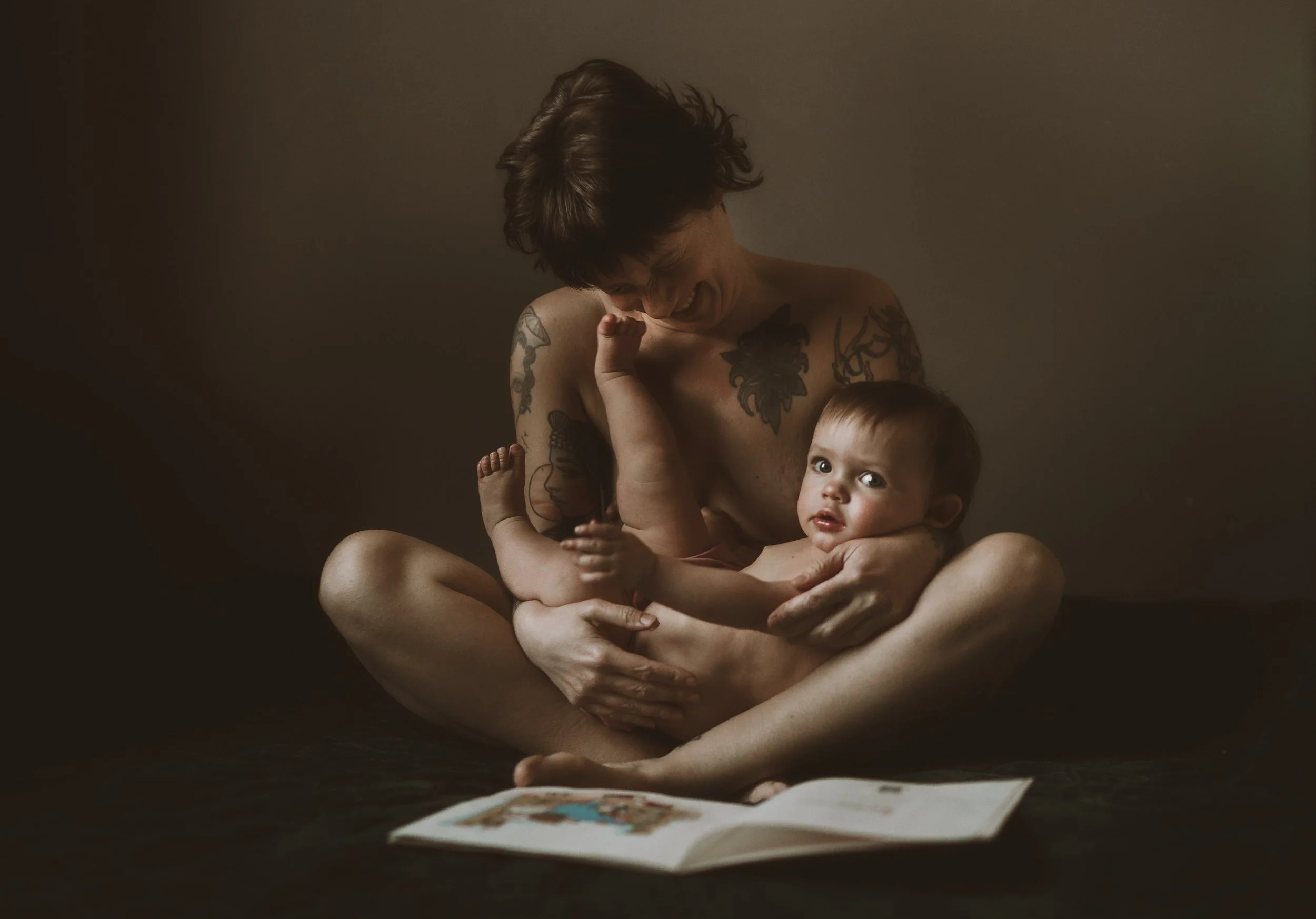 A woman with tattoos holding a baby, sitting cross-legged, with a paper or book open in front of them.