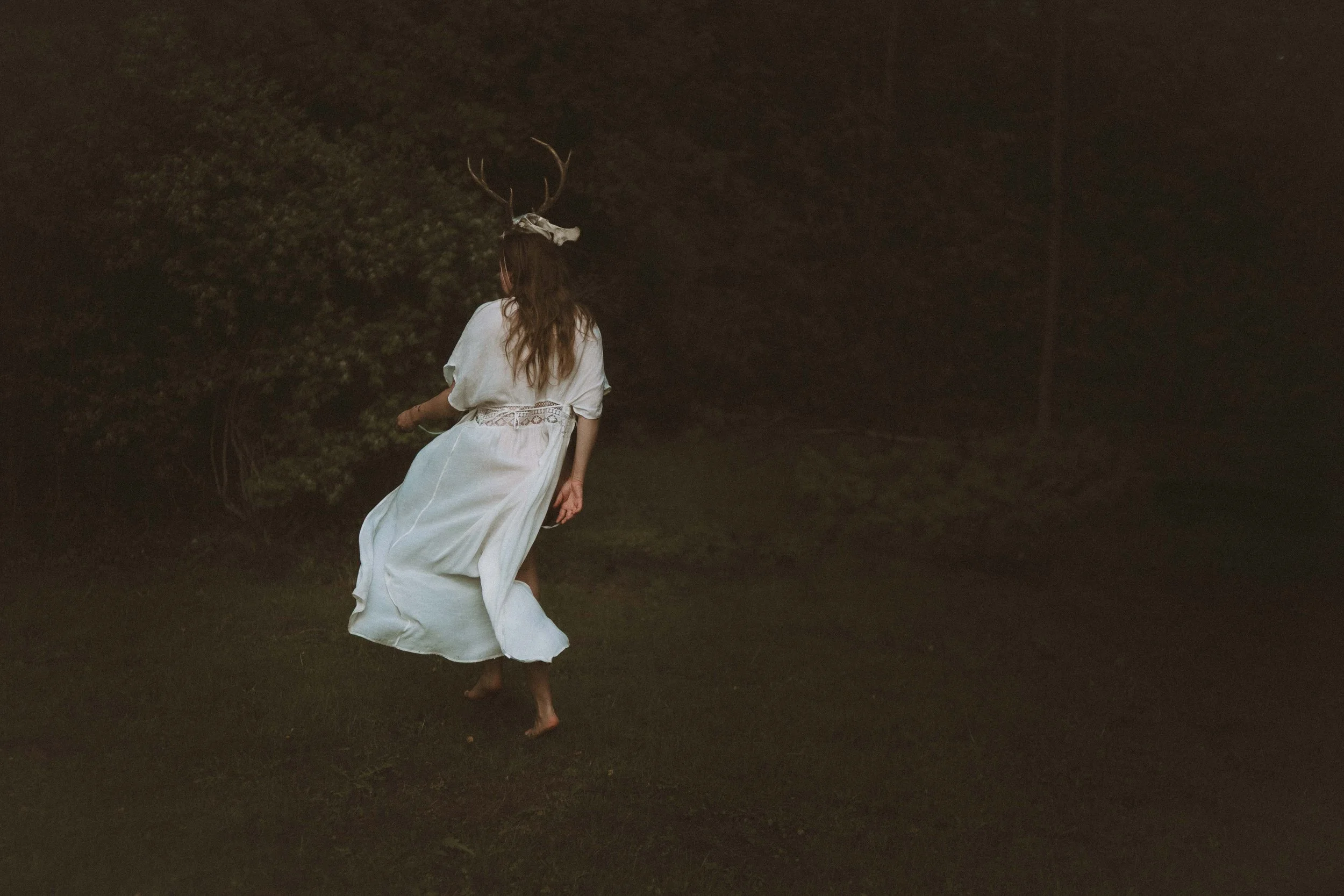 A woman wearing a white dress with antlers on her head is walking barefoot in a dark outdoor area at night.