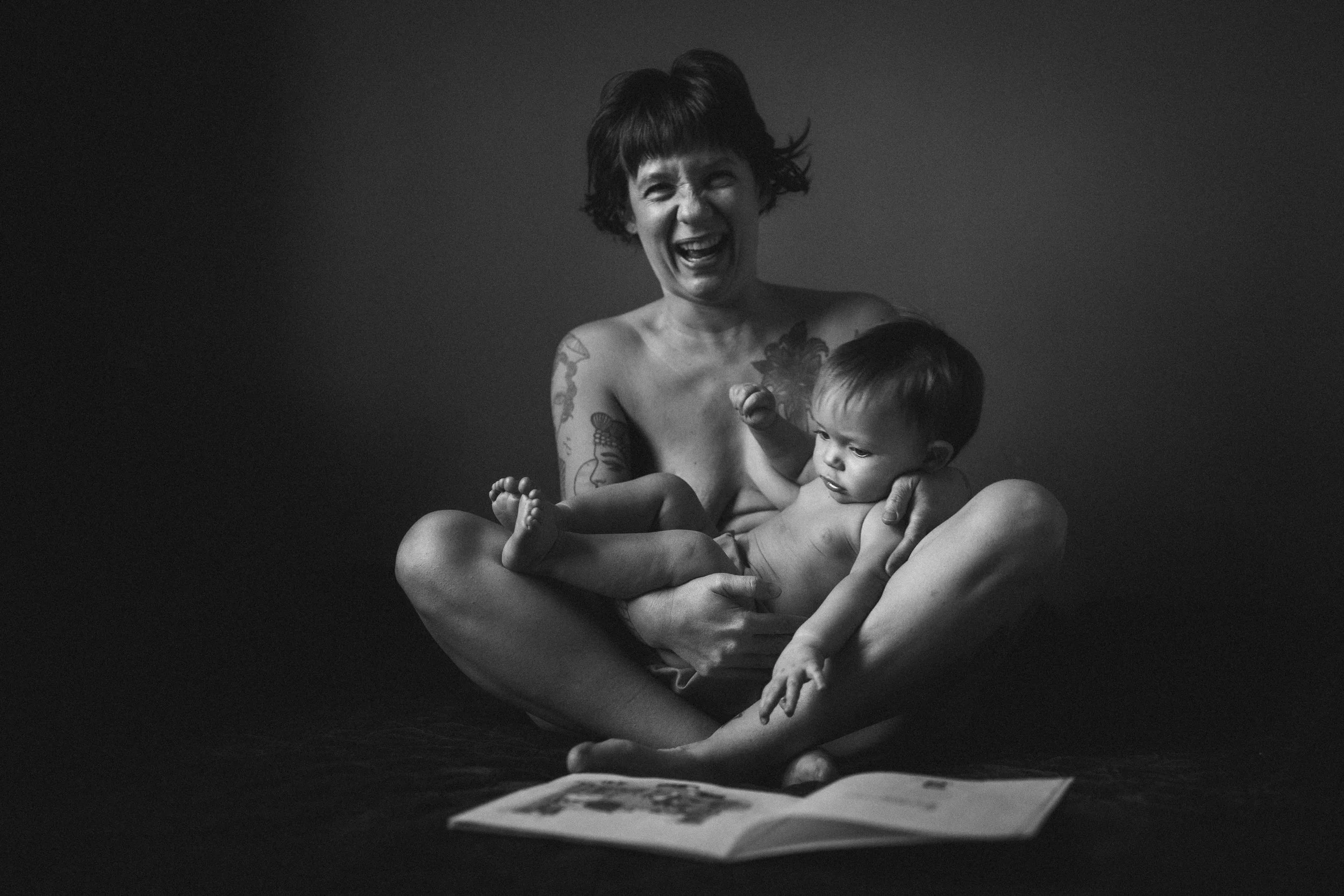 Black and white photo of a joyful woman with tattoos holding a young child while sitting on the floor, with a book open in front of them.