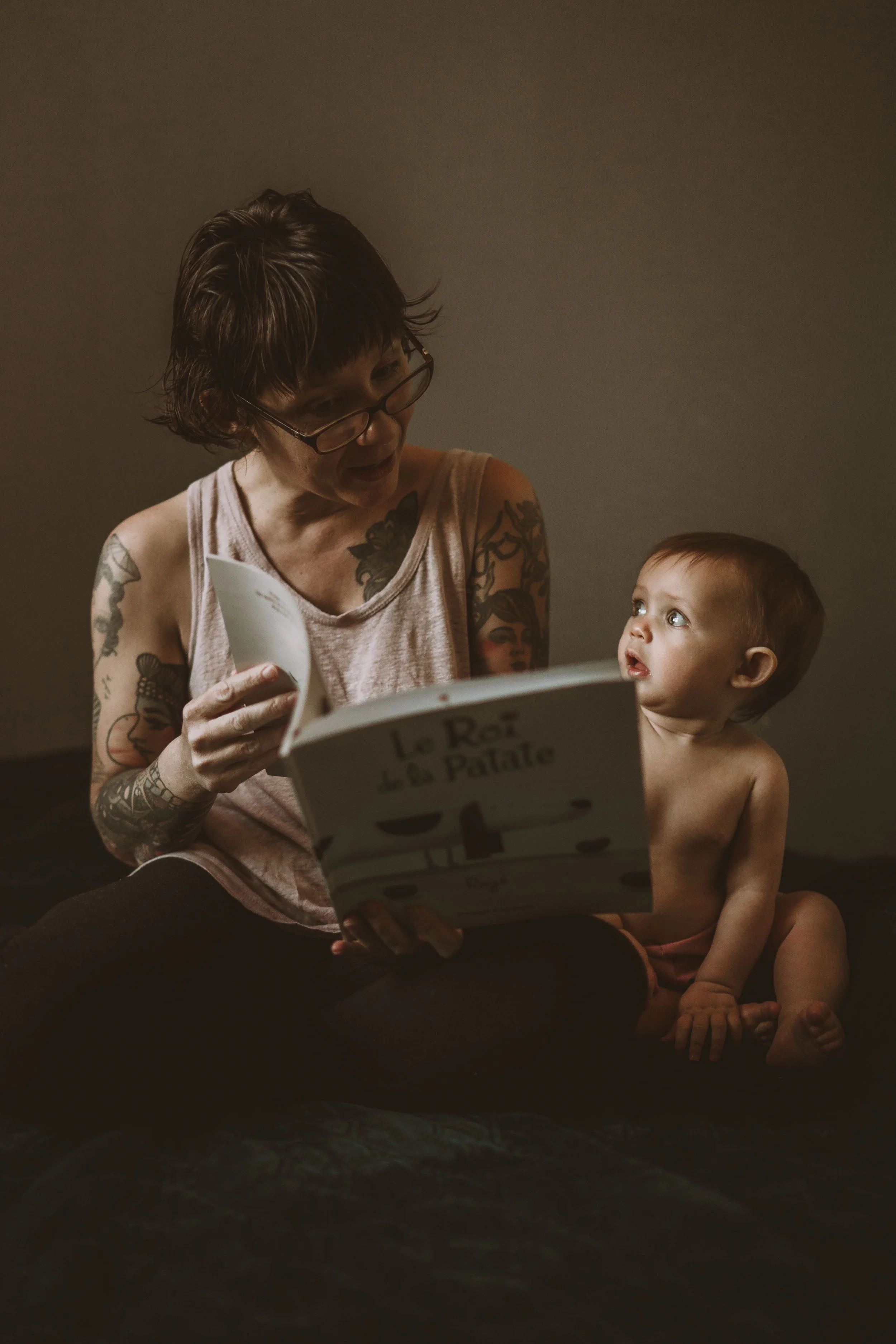 A woman with tattoos and glasses sitting and reading a book to a young child, who is sitting next to her shirtless. The woman has short, dark hair and is focused on the book, while the child's gaze is directed upward at her with curiosity. The backgr