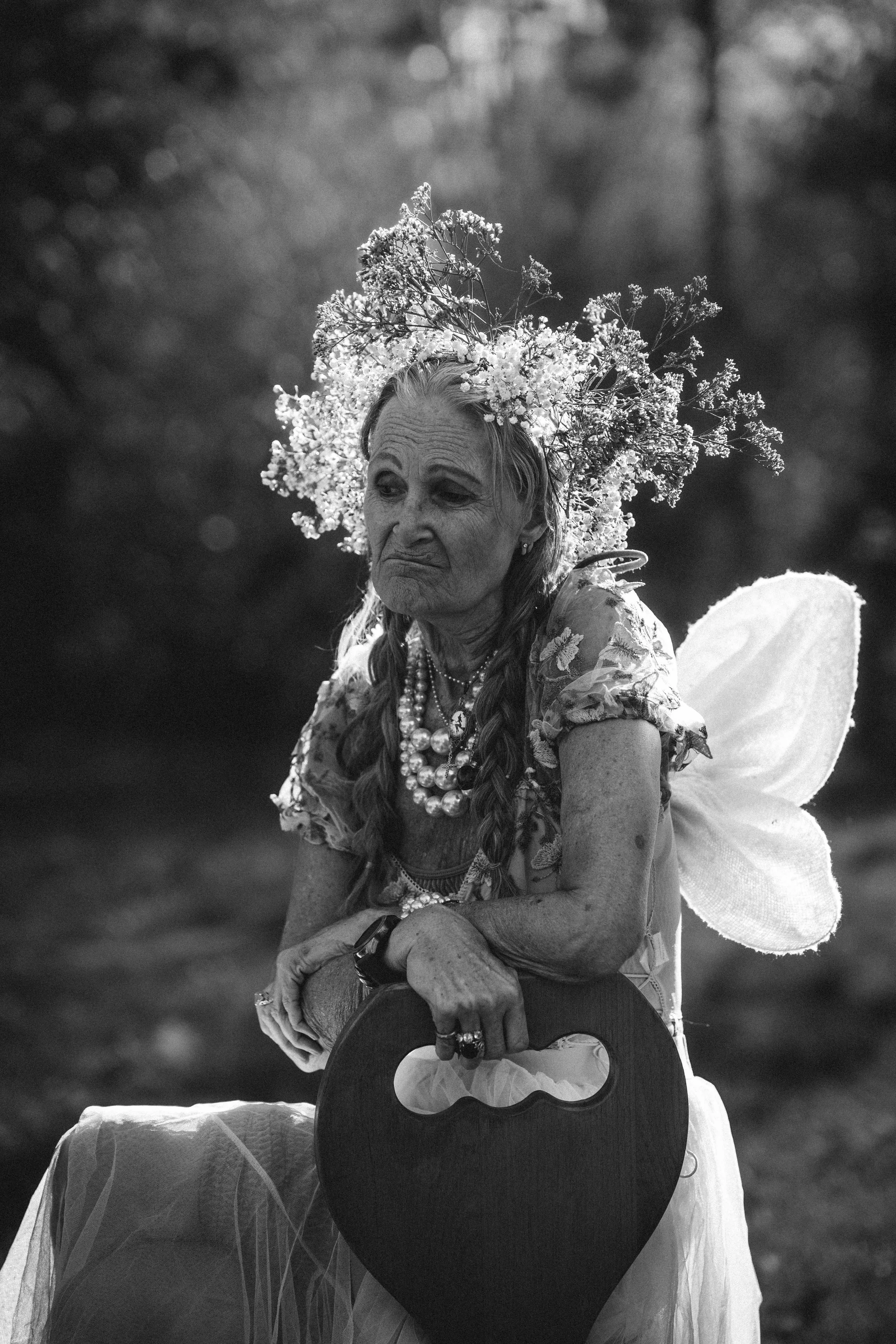 A woman dressed as a fairy with a floral crown, fairy wings, and jewelry, sitting outdoors in a black and white photograph with a blurred background.