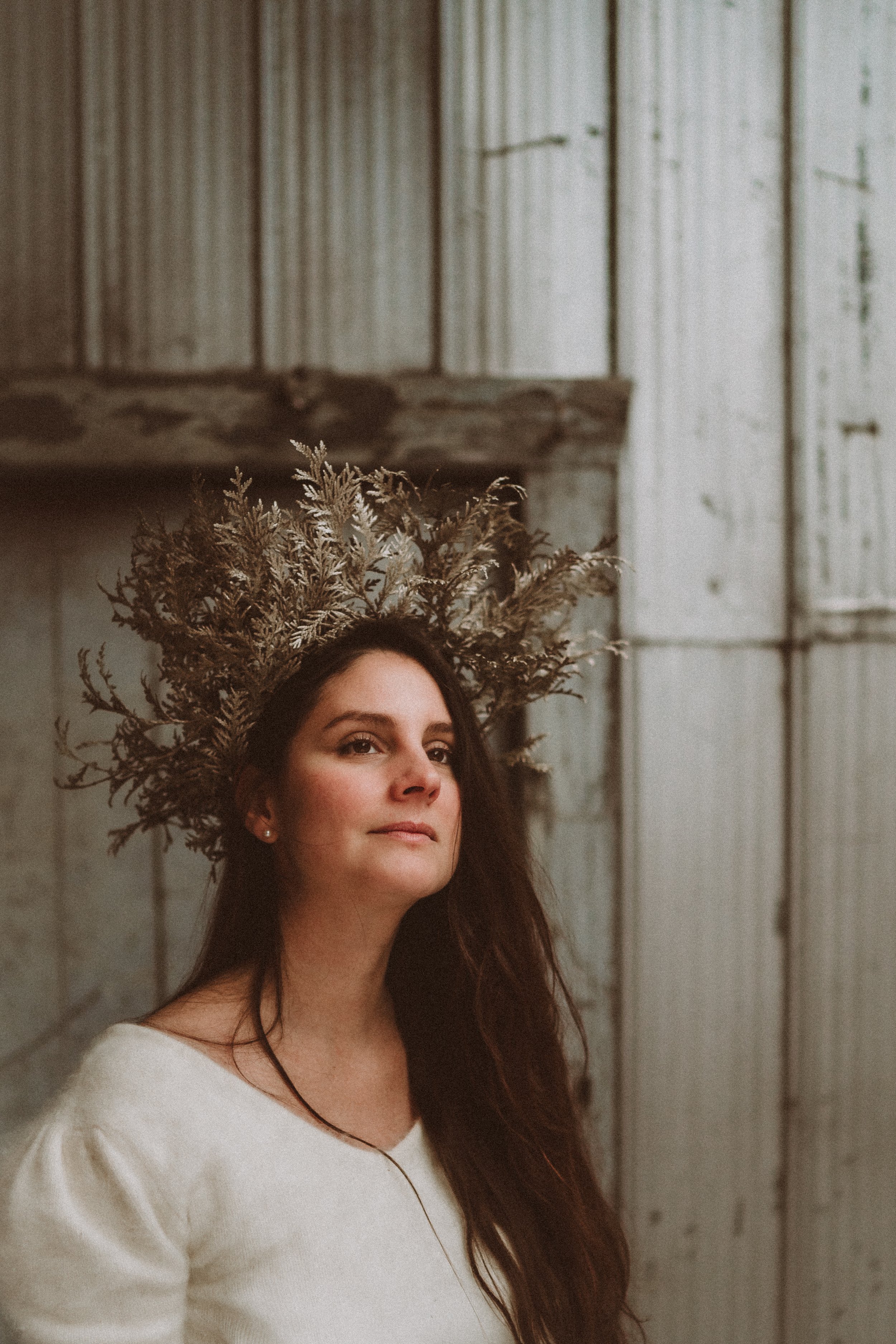 A woman with long dark hair wearing a cream-colored top, adorned with a crown made of dried plants or foliage, standing against a rustic, weathered wooden background.