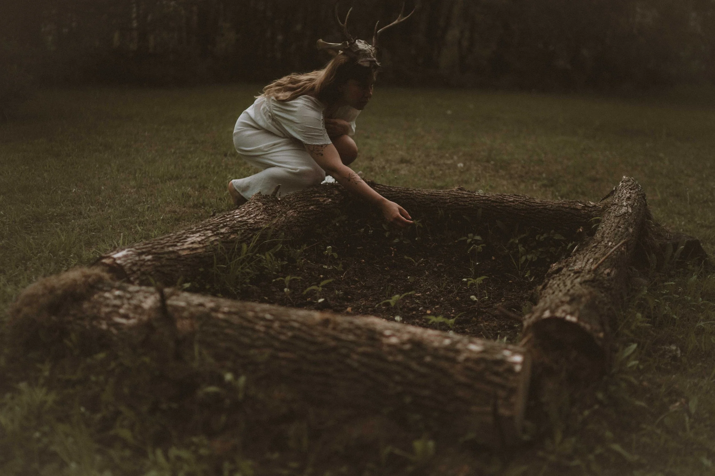 Person wearing a deer mask, squatting on the ground in a wooded area, with a large fallen tree trunk in front.