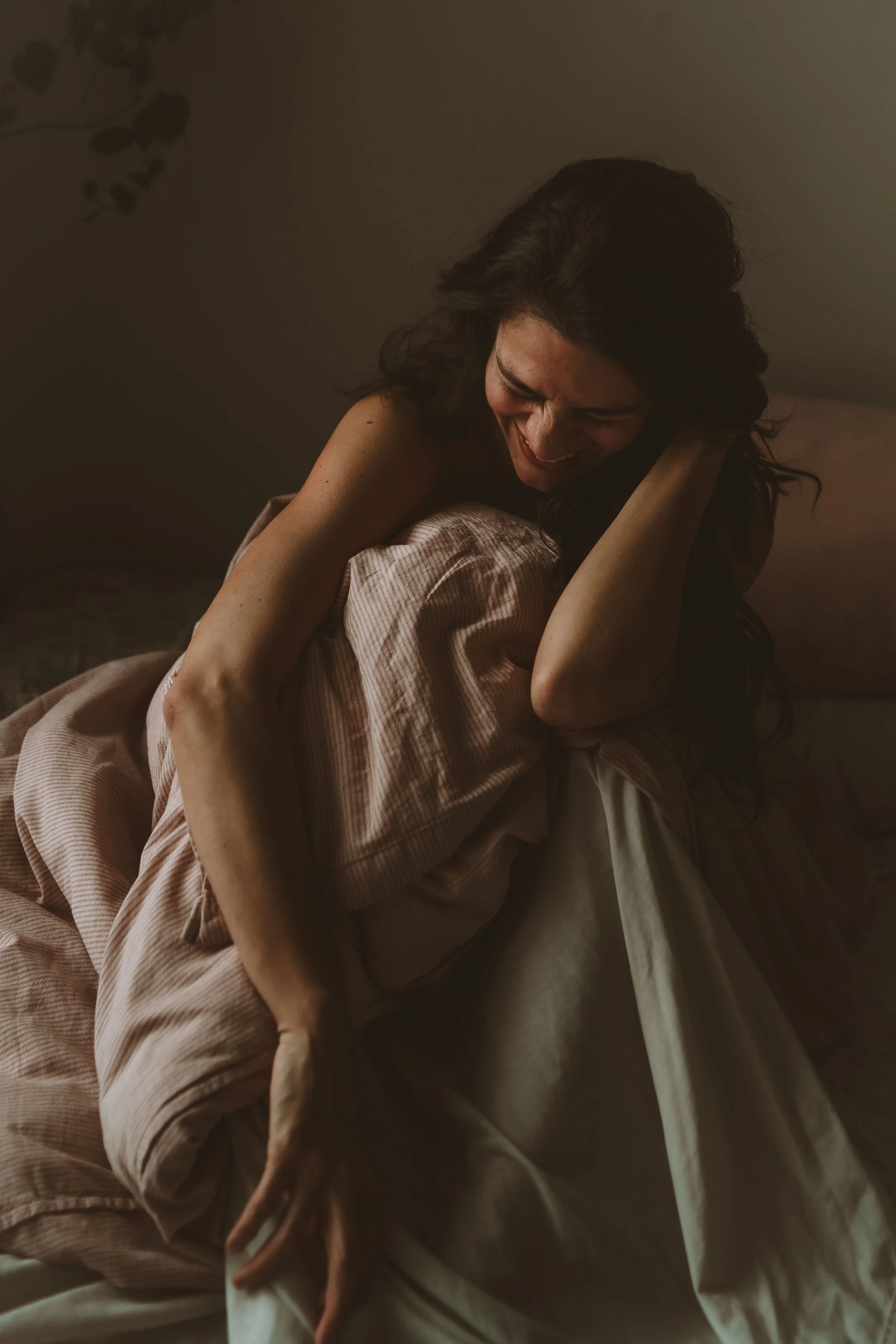 A woman with dark hair, smiling and with eyes closed, is sitting on a bed in soft, warm lighting, wearing a pink striped shirt and hugging herself.