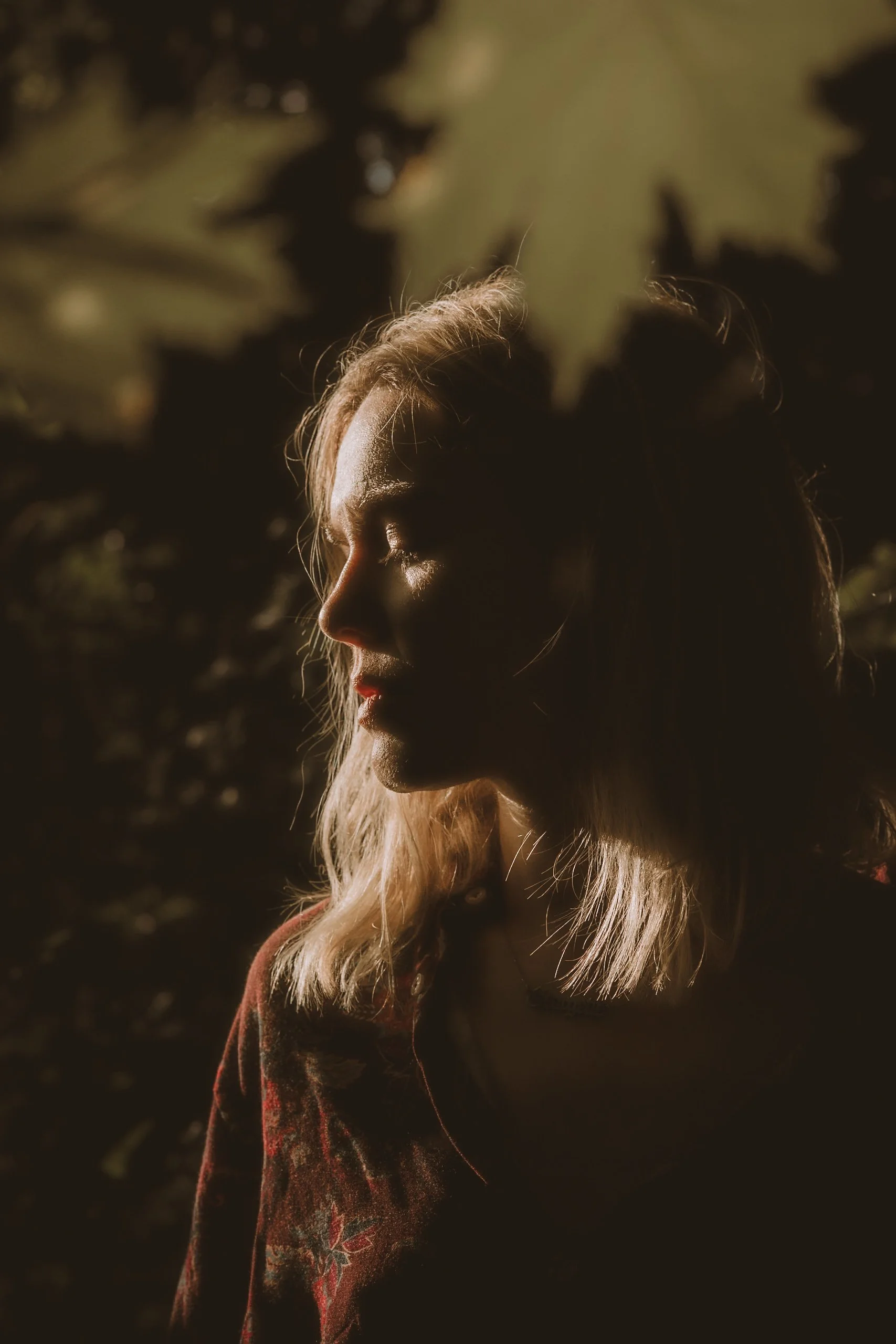 A woman with closed eyes and long, light-colored hair standing outdoors with sunlight highlighting her face and hair, surrounded by dark leafy trees.