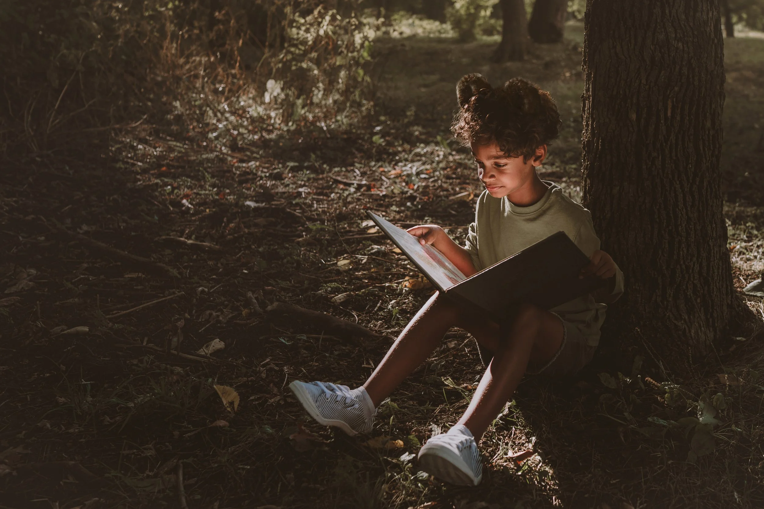 A young girl with curly hair tied in pigtails, sitting against a tree trunk in a forest, reading a book, with sunlight filtering through the trees.