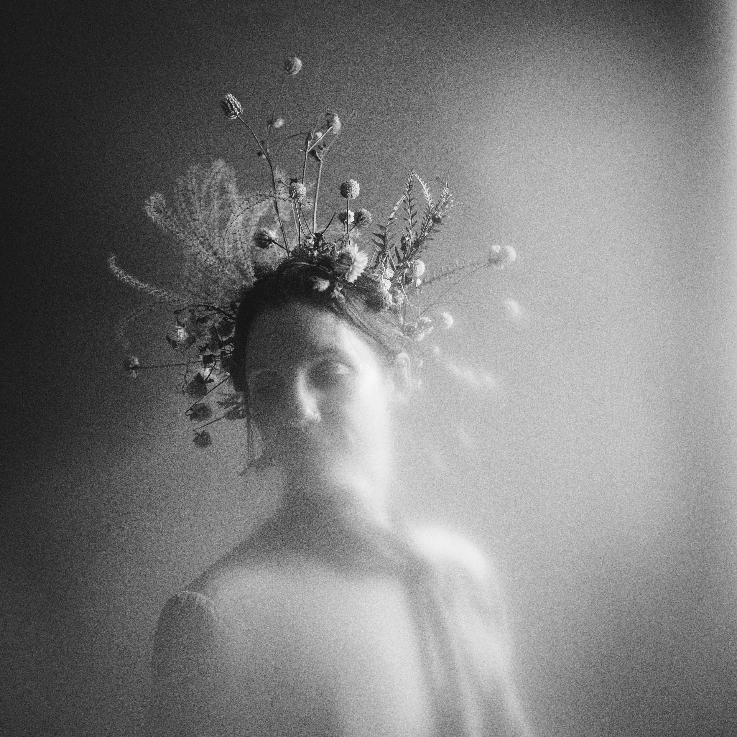 A black-and-white portrait of a woman with a floral crown on her head, looking slightly downward with her eyes closed against a plain background.