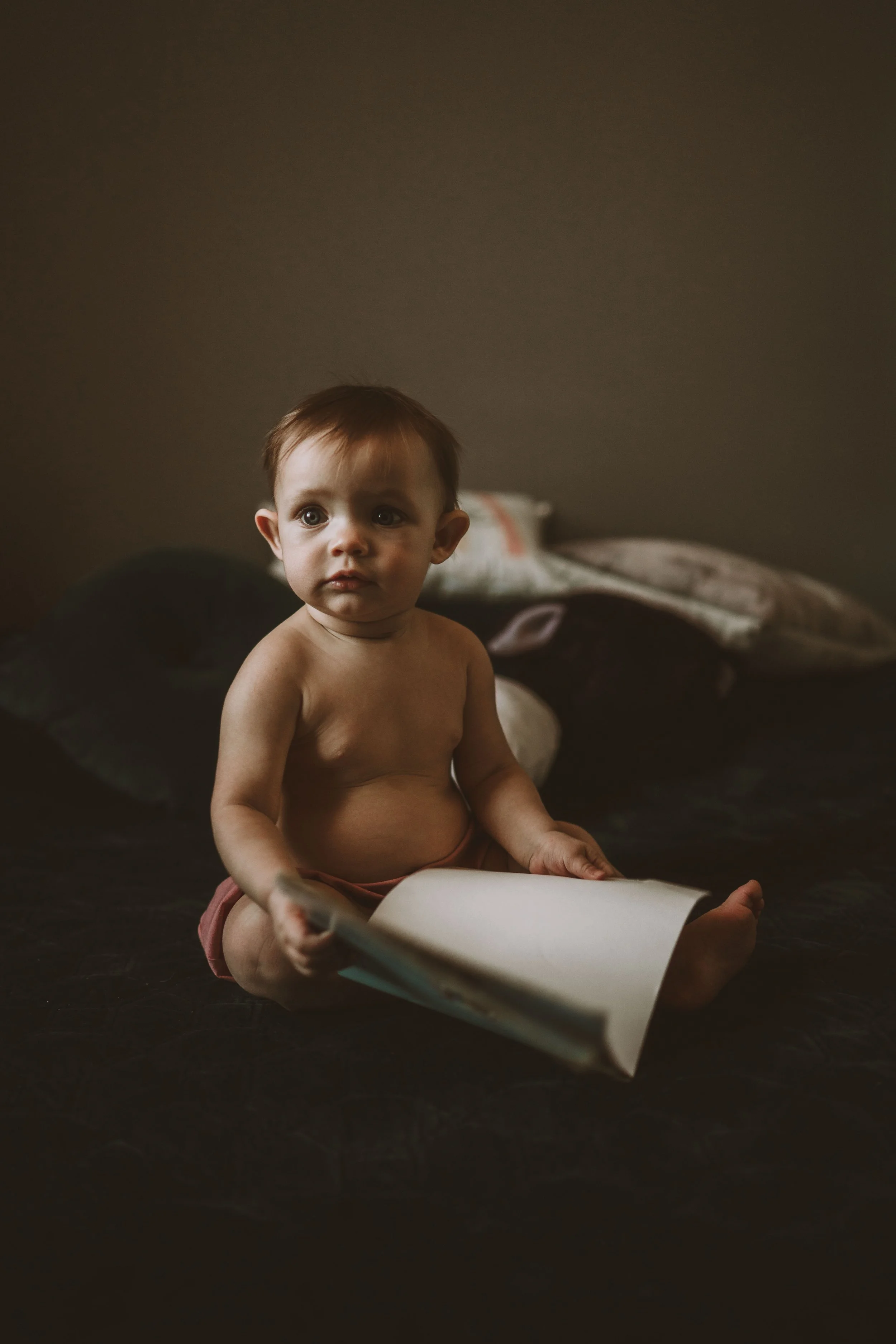 A shirtless young boy with brown hair sits on a dark surface, holding a piece of paper or a book, with a somber expression. In the background, there are several pillows on a bed or couch against a plain wall.