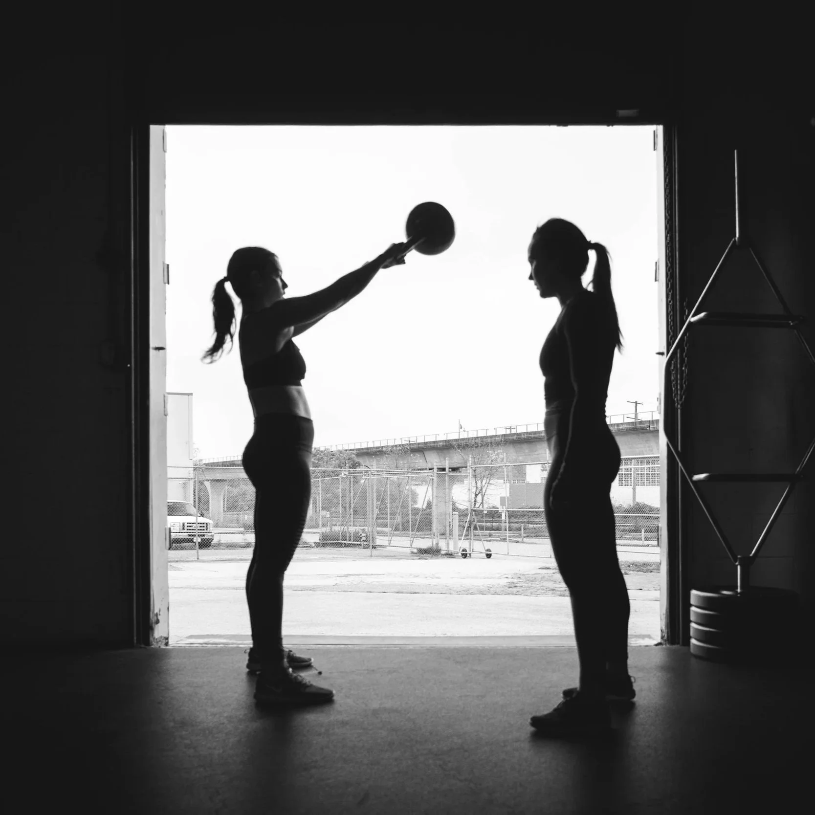 Silhouettes of two women in sportswear standing face to face inside a gym, one holding a medicine ball over her head, with a baseball field and stadium in the background outside the open gym door.
