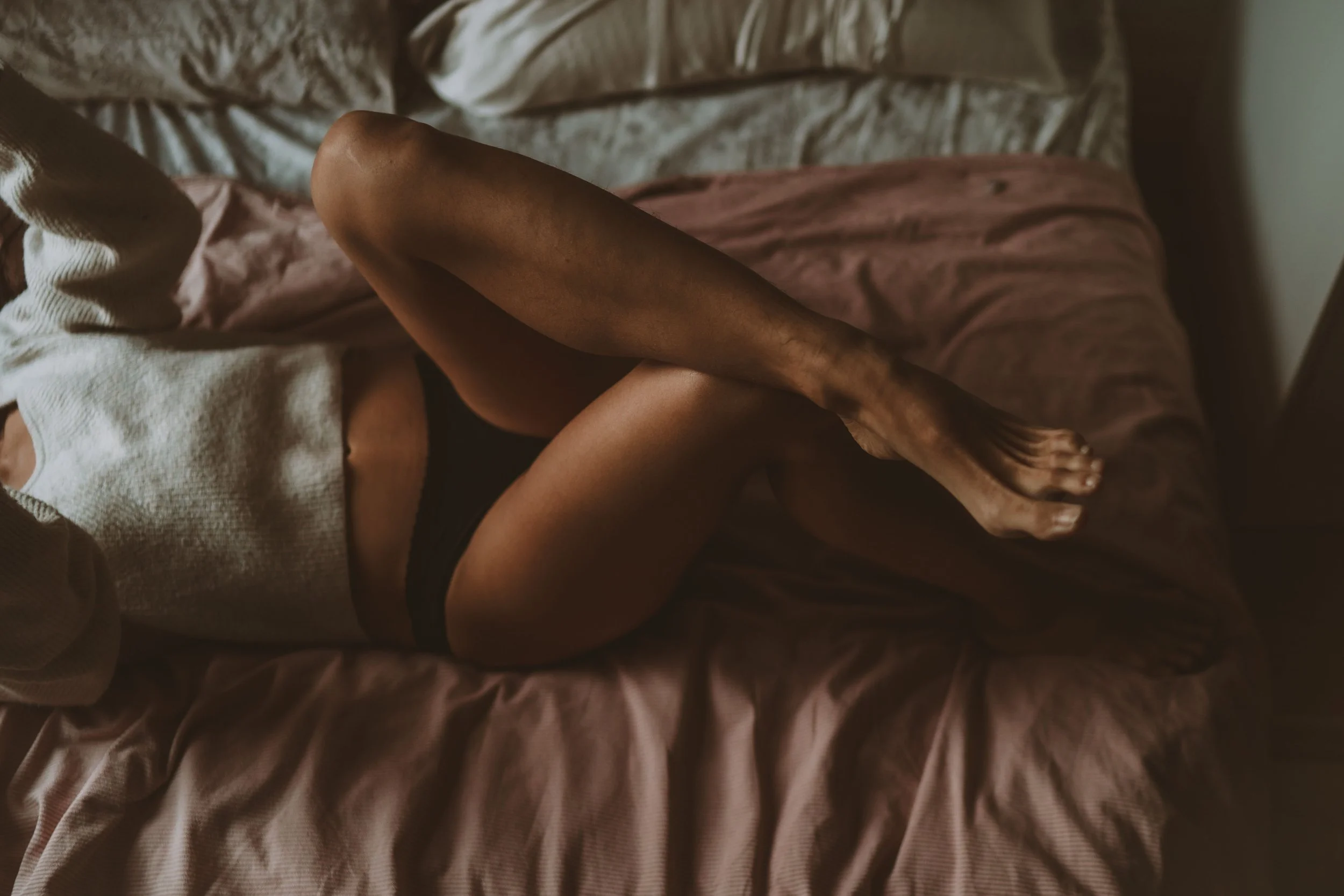 A person lying on a bed with their legs crossed at the ankles, wearing black underwear and a beige sweater, on pink and beige sheets.