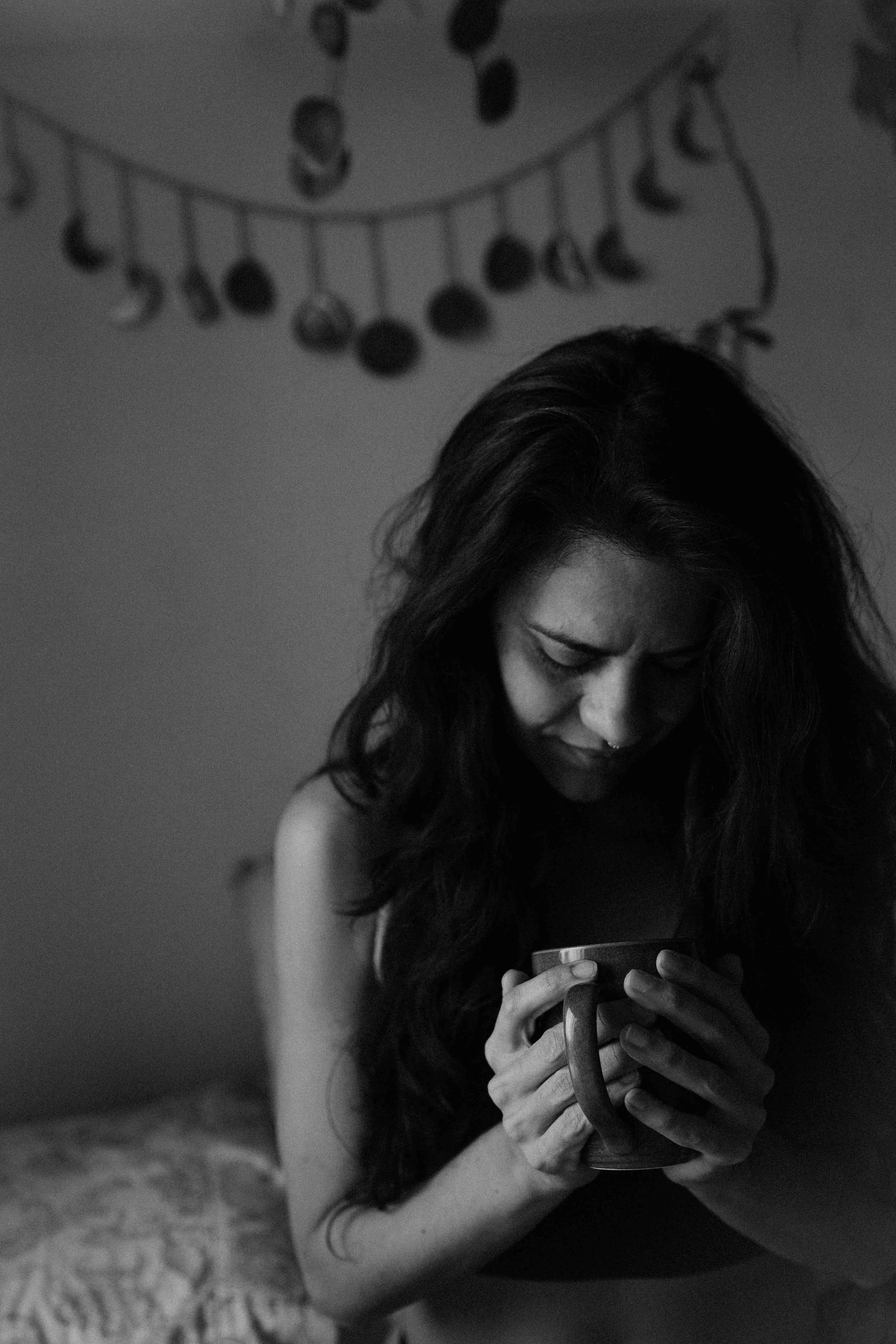 A black-and-white photo of a woman with long, wavy hair holding a mug, looking down with a contemplative expression, in a dimly lit room with a decorative curtain or string of objects hanging in the background.