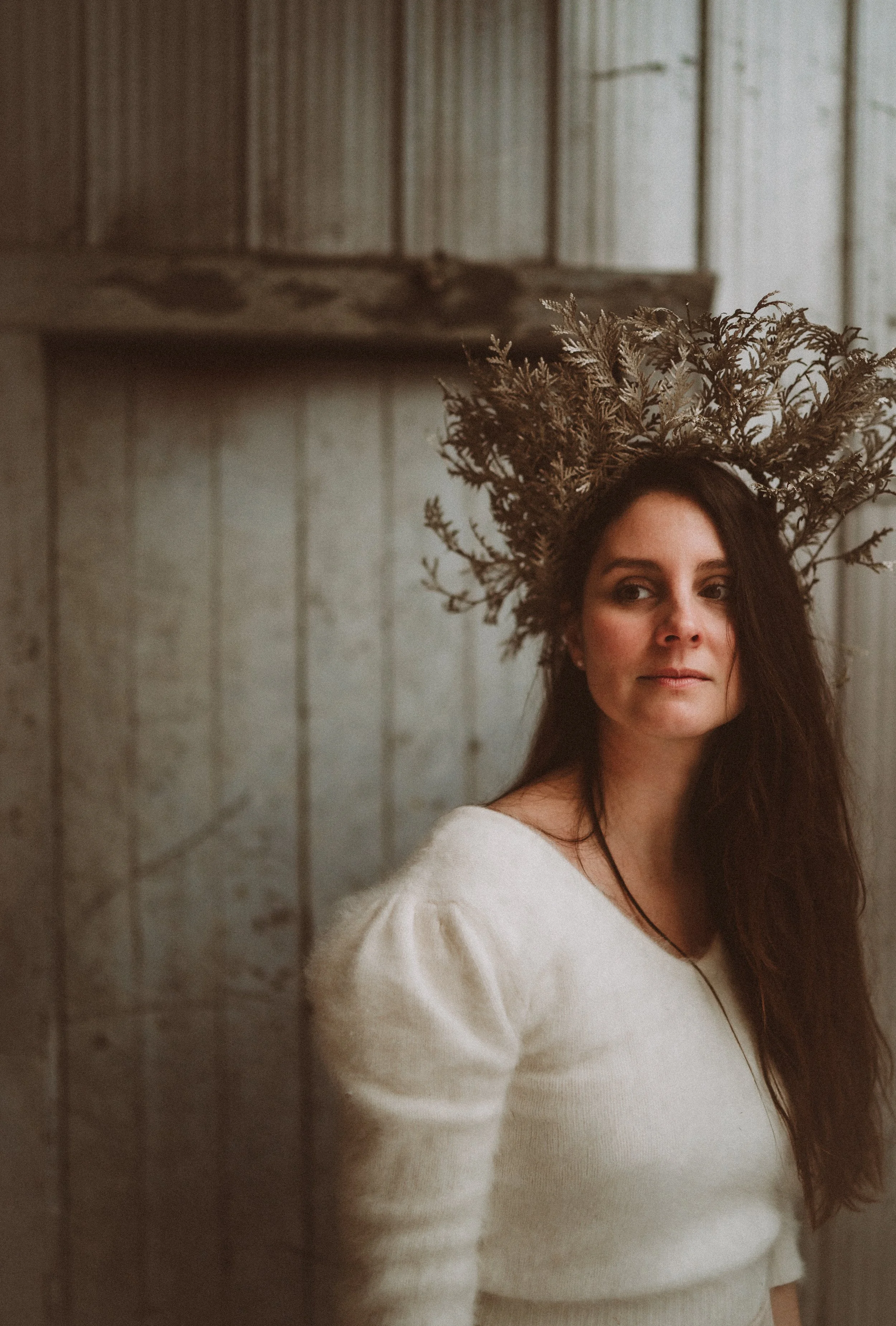 A woman with long dark hair wearing a white sweater and a crown made of dried branches standing against a wooden wall.