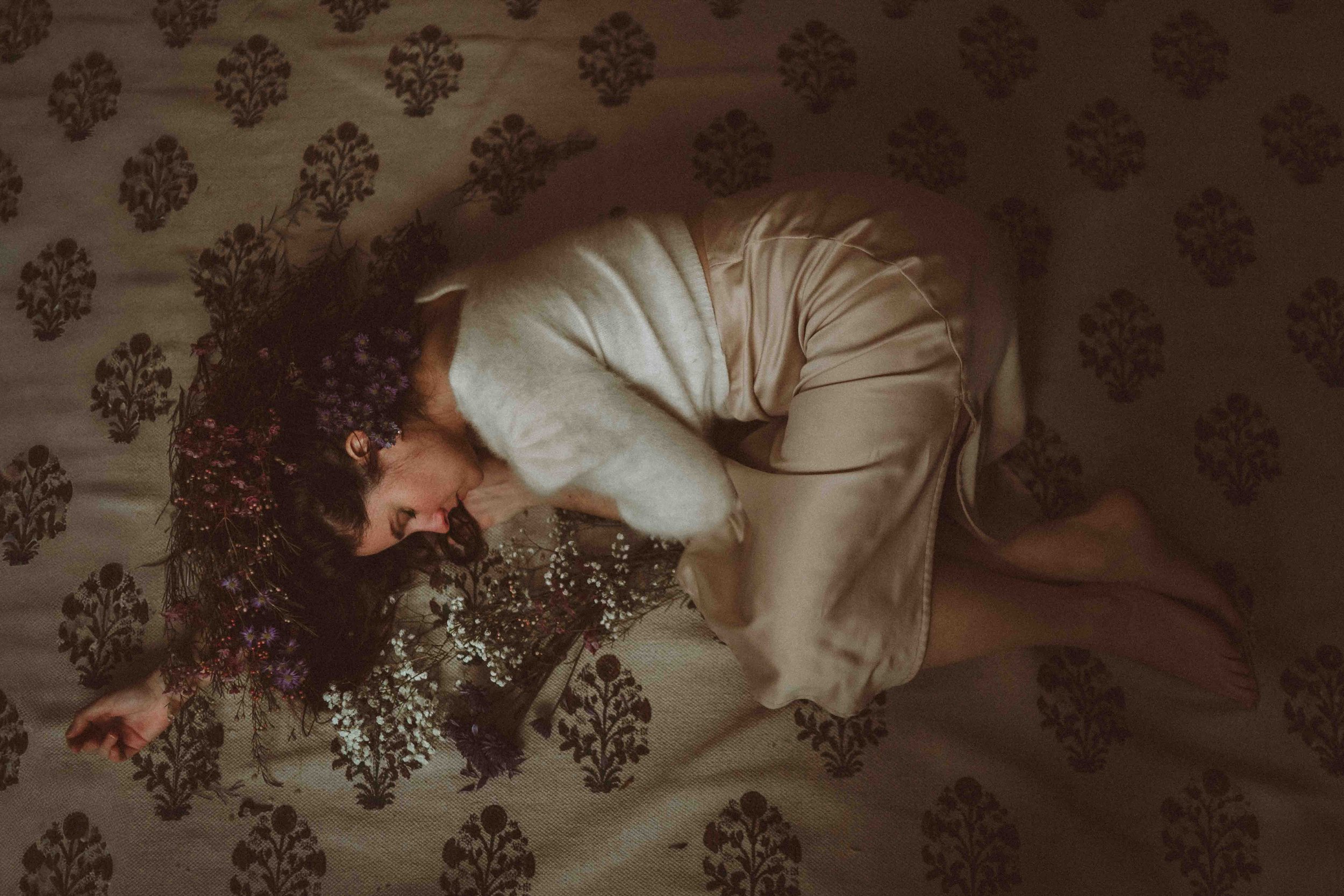 A woman with dark hair and a floral crown is lying on her side on a patterned fabric surface, wearing a beige satin nightgown and pink slippers, with her eyes closed.