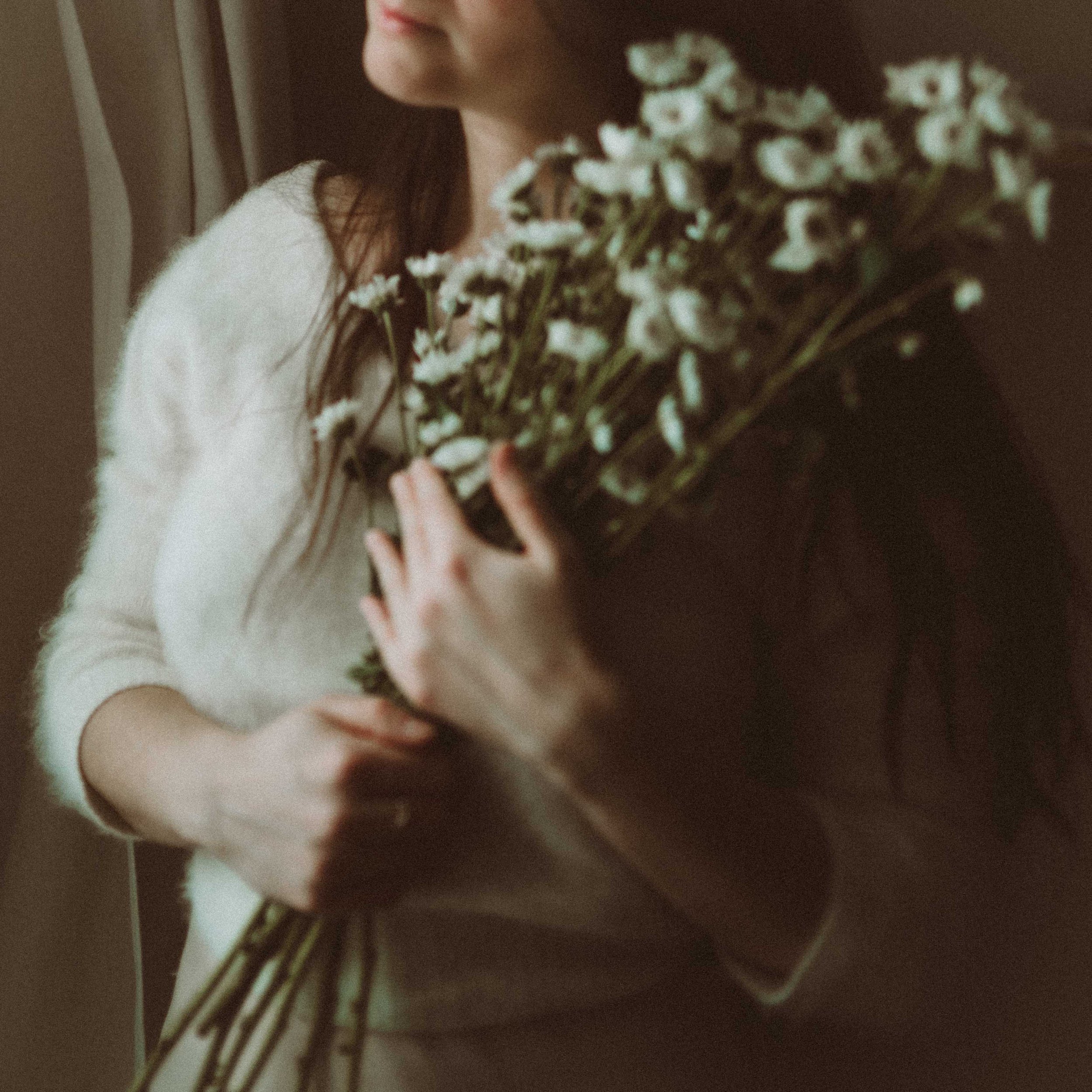A woman holding a large bouquet of white flowers near her chest, wearing a light-colored sweater, with a blurred background.