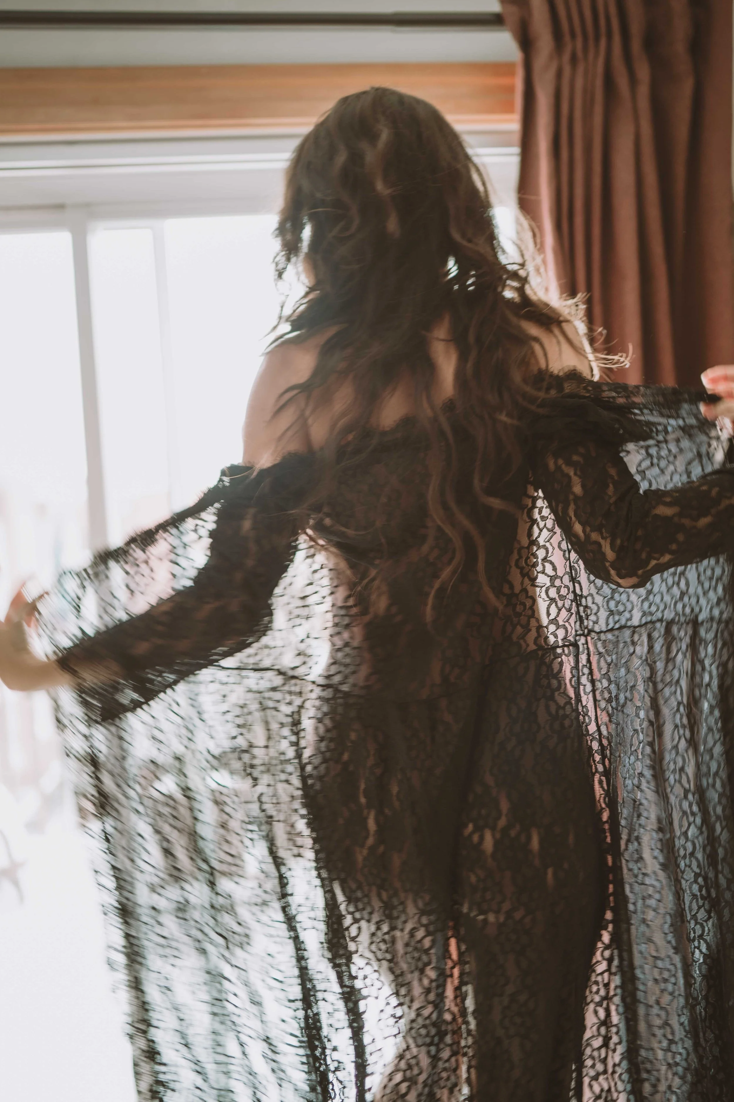 A woman in a black lace dress standing near a window, brushing her hair back, with sunlight shining through the window.