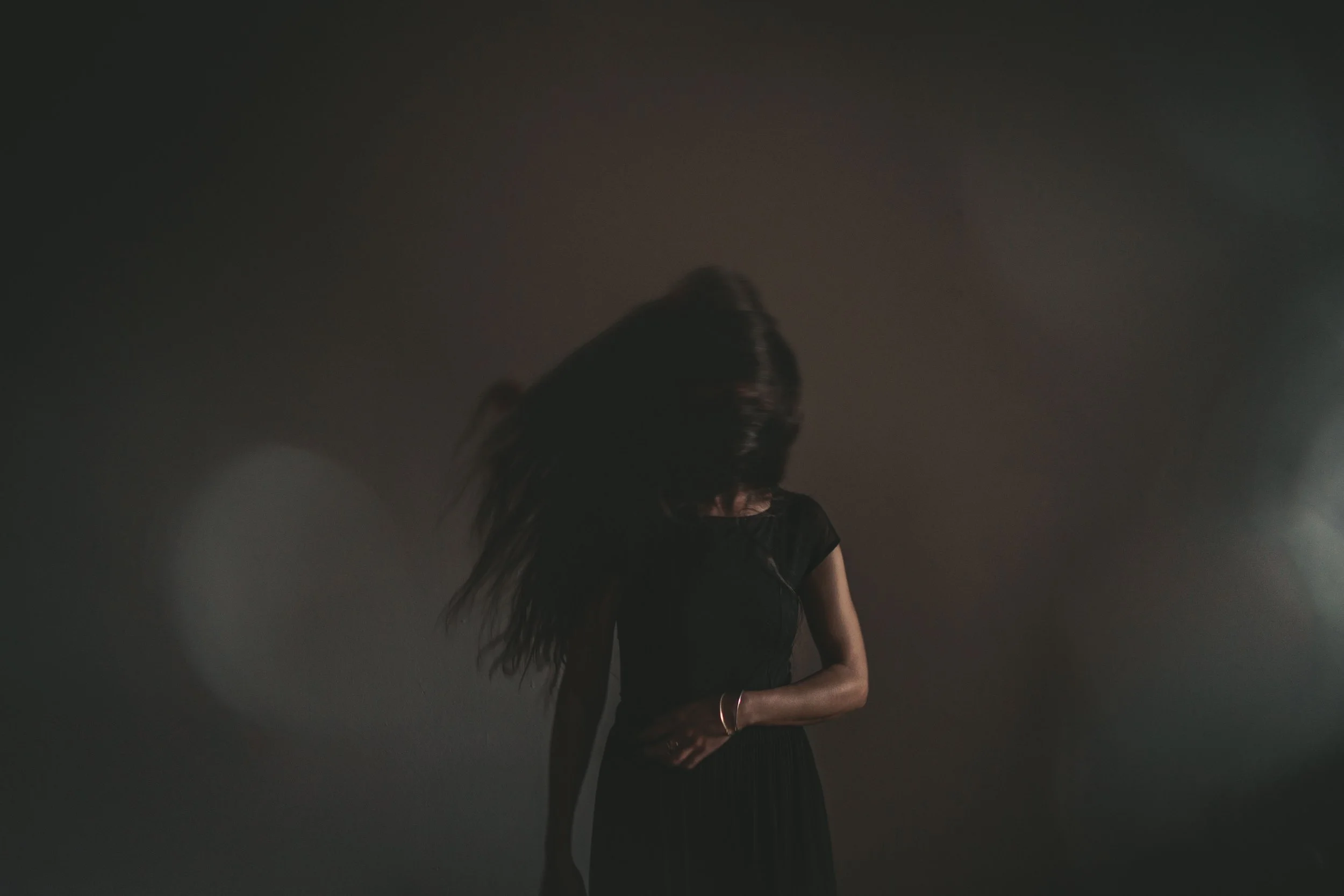 A woman with long dark hair wearing a black dress, looking down with her hair covering her face, in a dimly lit setting with soft light and dark background.