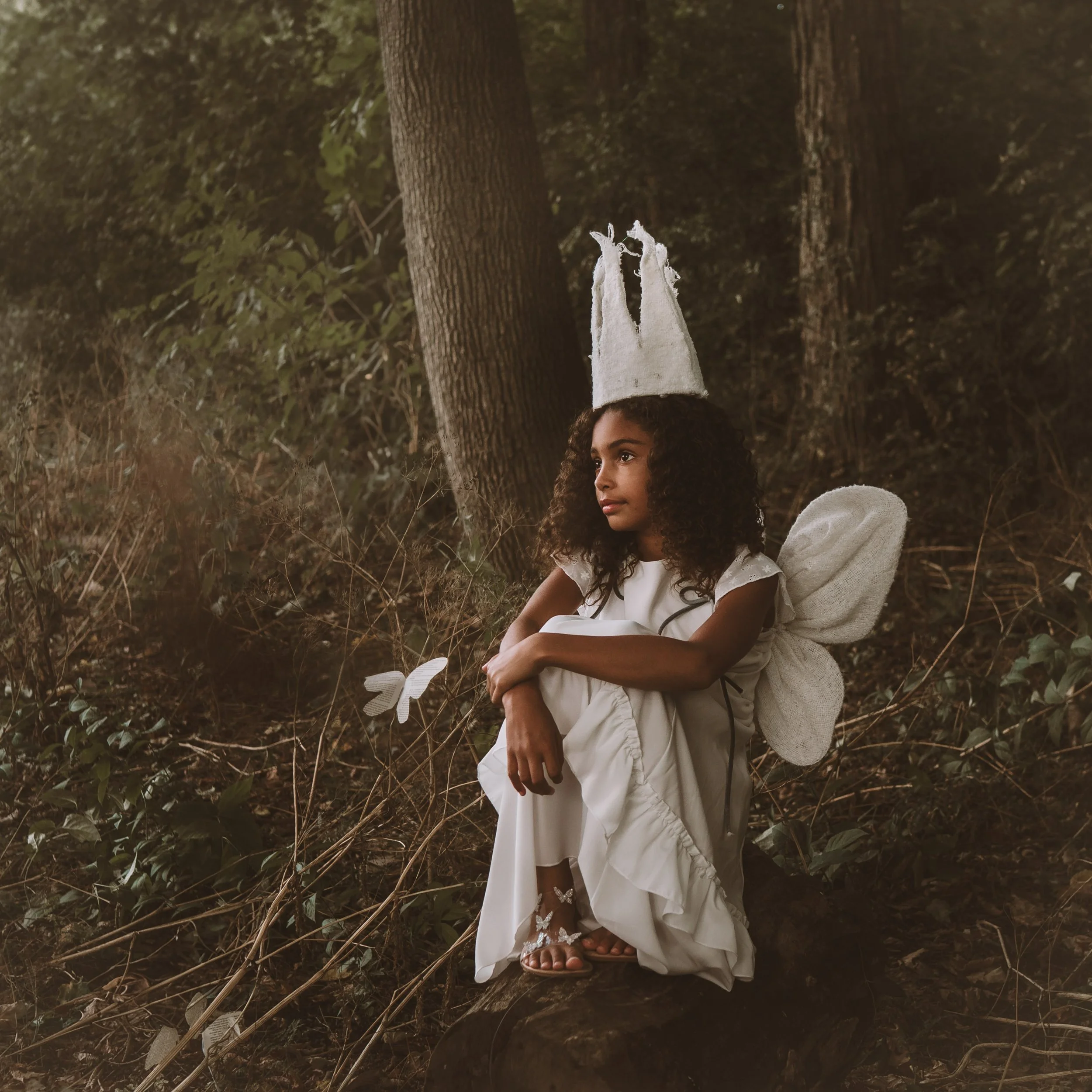 A young girl with curly hair sitting outdoors among trees and foliage, wearing a white dress and sandals, with a white cloth on her head and a white nightingale perched on her finger.
