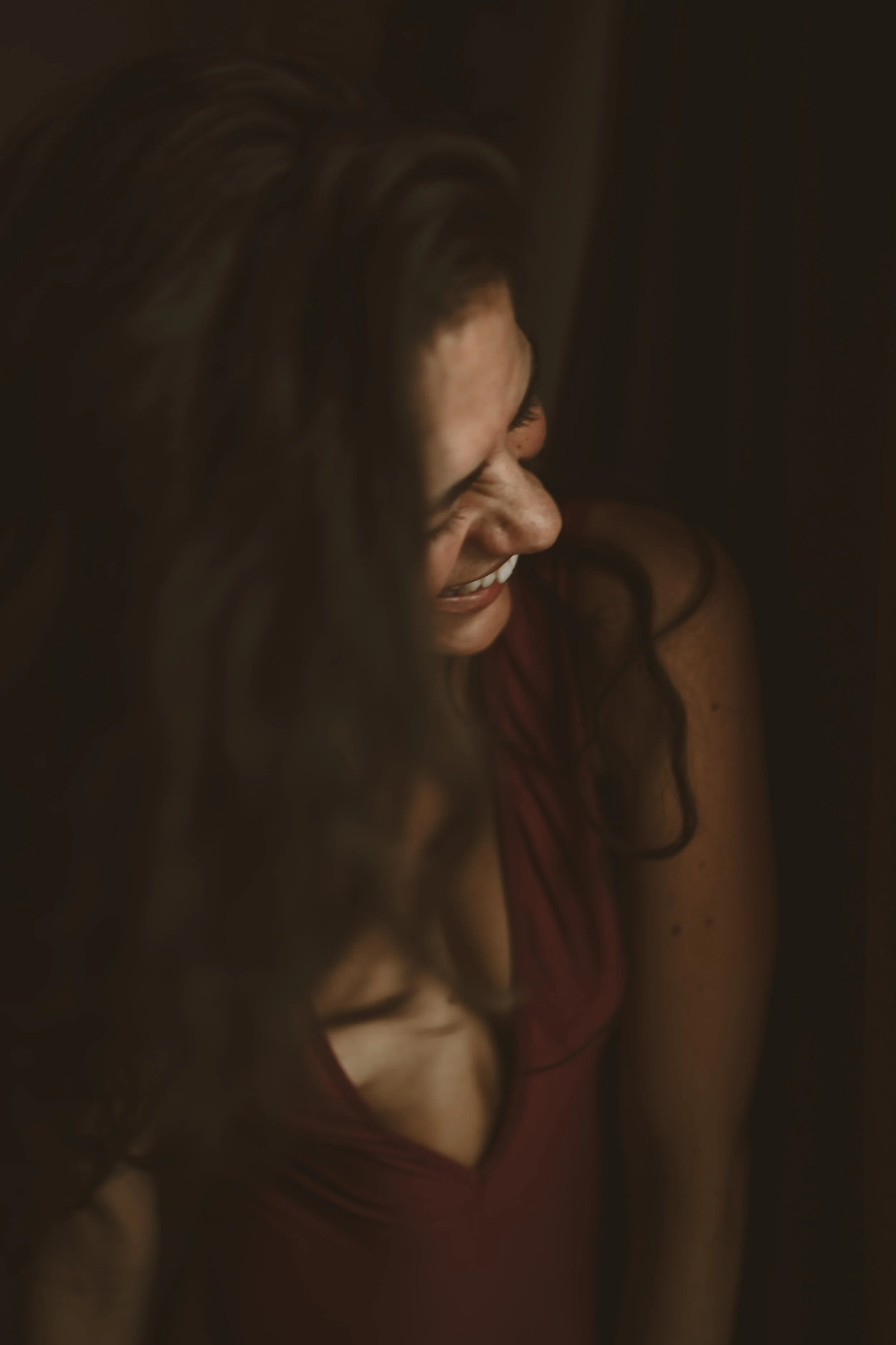 A woman with long wavy hair smiling and looking down in a dark setting, wearing a burgundy dress with a plunging neckline.
