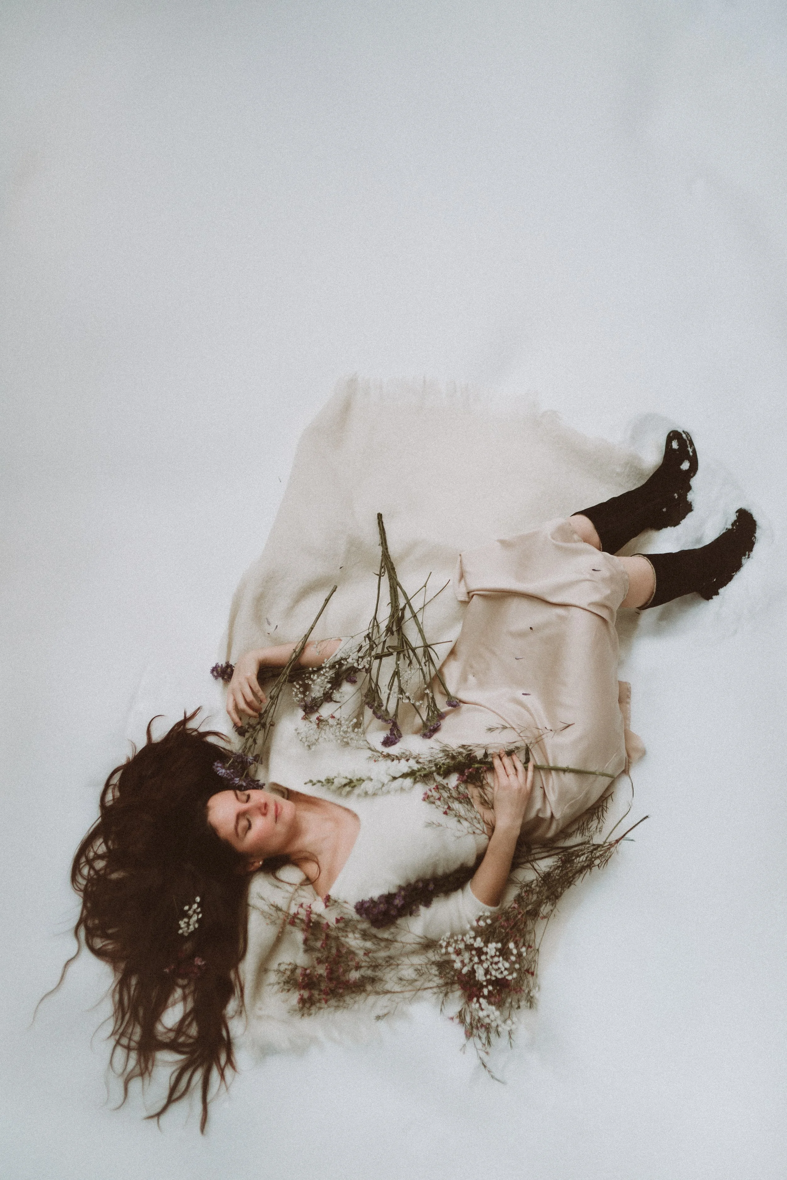 A woman lying on the floor surrounded by dried flowers, wearing a cream-colored dress and black socks.