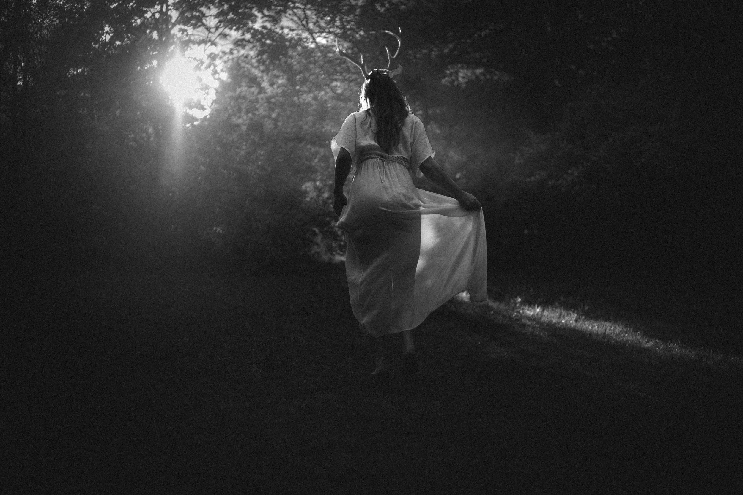 A black and white photo of a person with long hair in a flowing dress, walking in a wooded area with sunlight filtering through the trees, wearing a deer antler headband.
